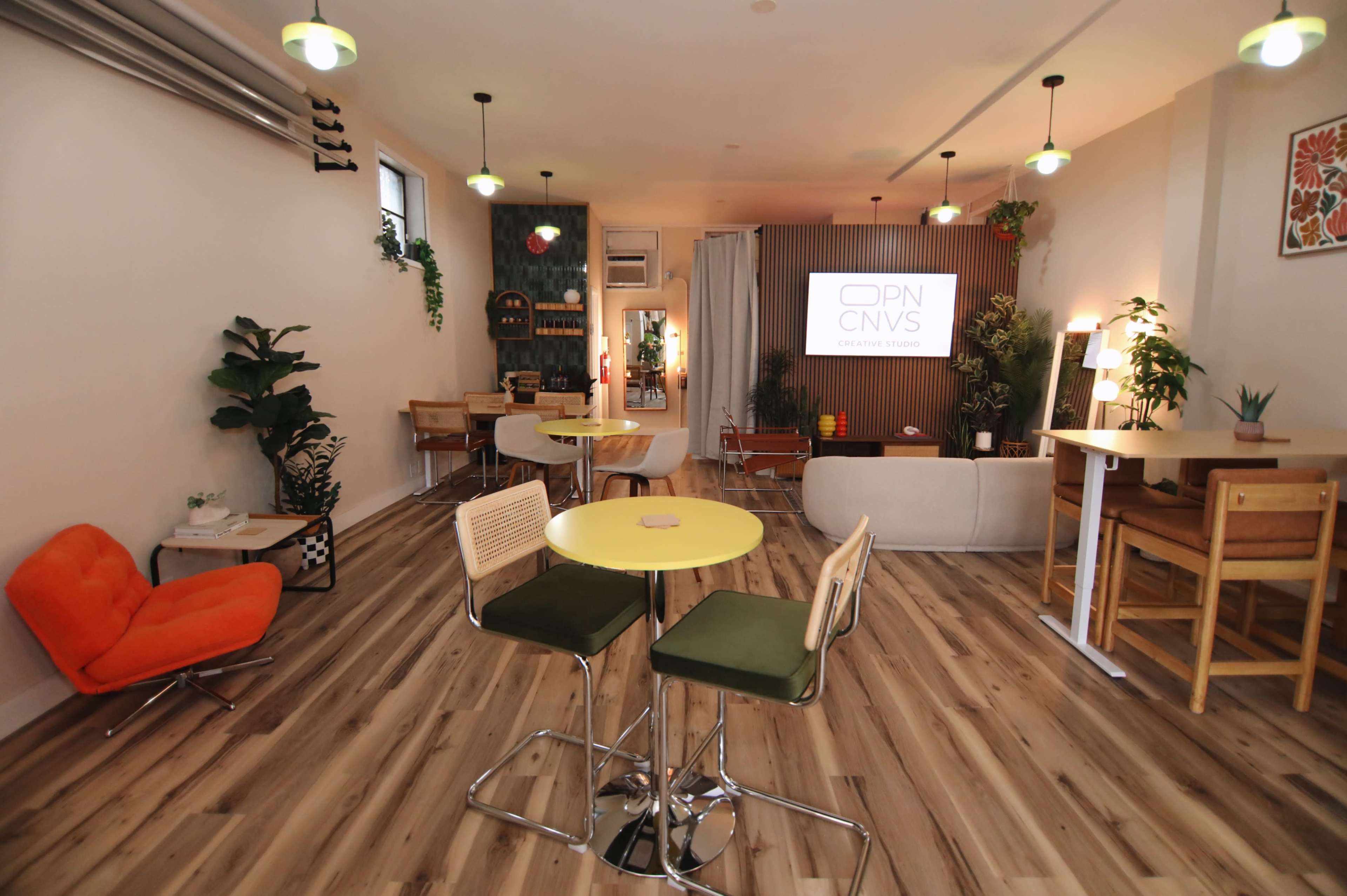 The image shows a modern co-working space featuring colorful furniture, plants, and a projector screen in a brightly lit environment.