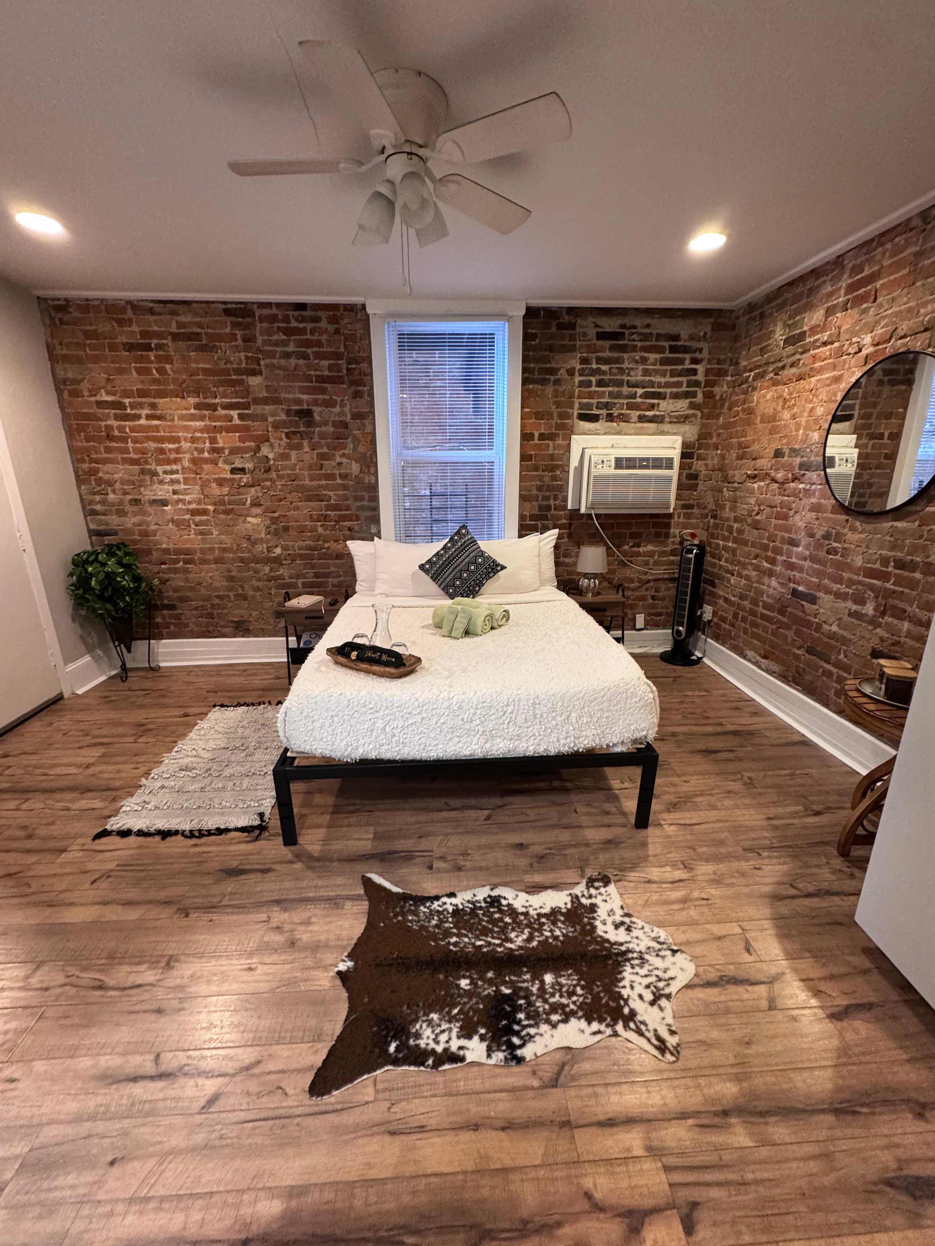 The image shows a bedroom featuring a bed against a brick wall, with wooden flooring, a small area rug, and an air conditioning unit on the wall.