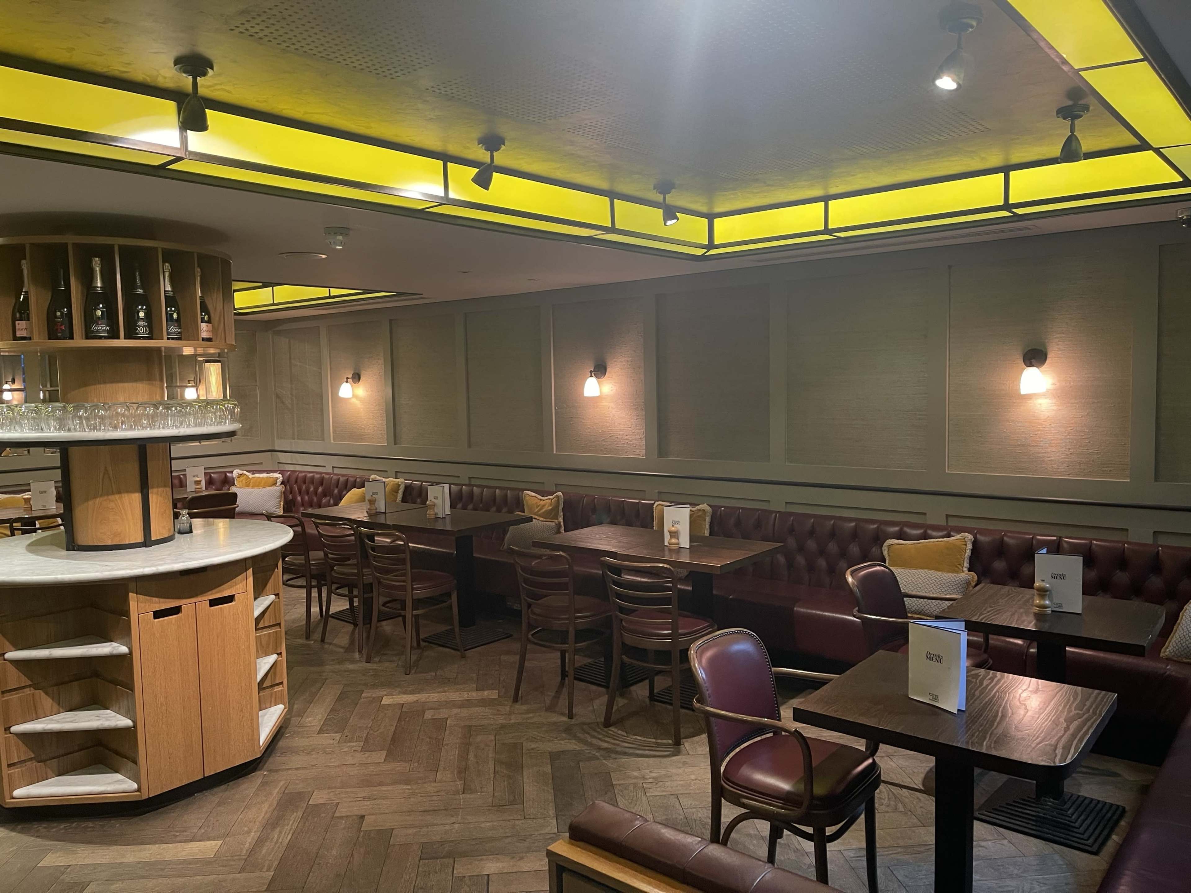 The image shows a casual dining area with dark wood tables and red leather seating, illuminated by yellow ceiling lights.