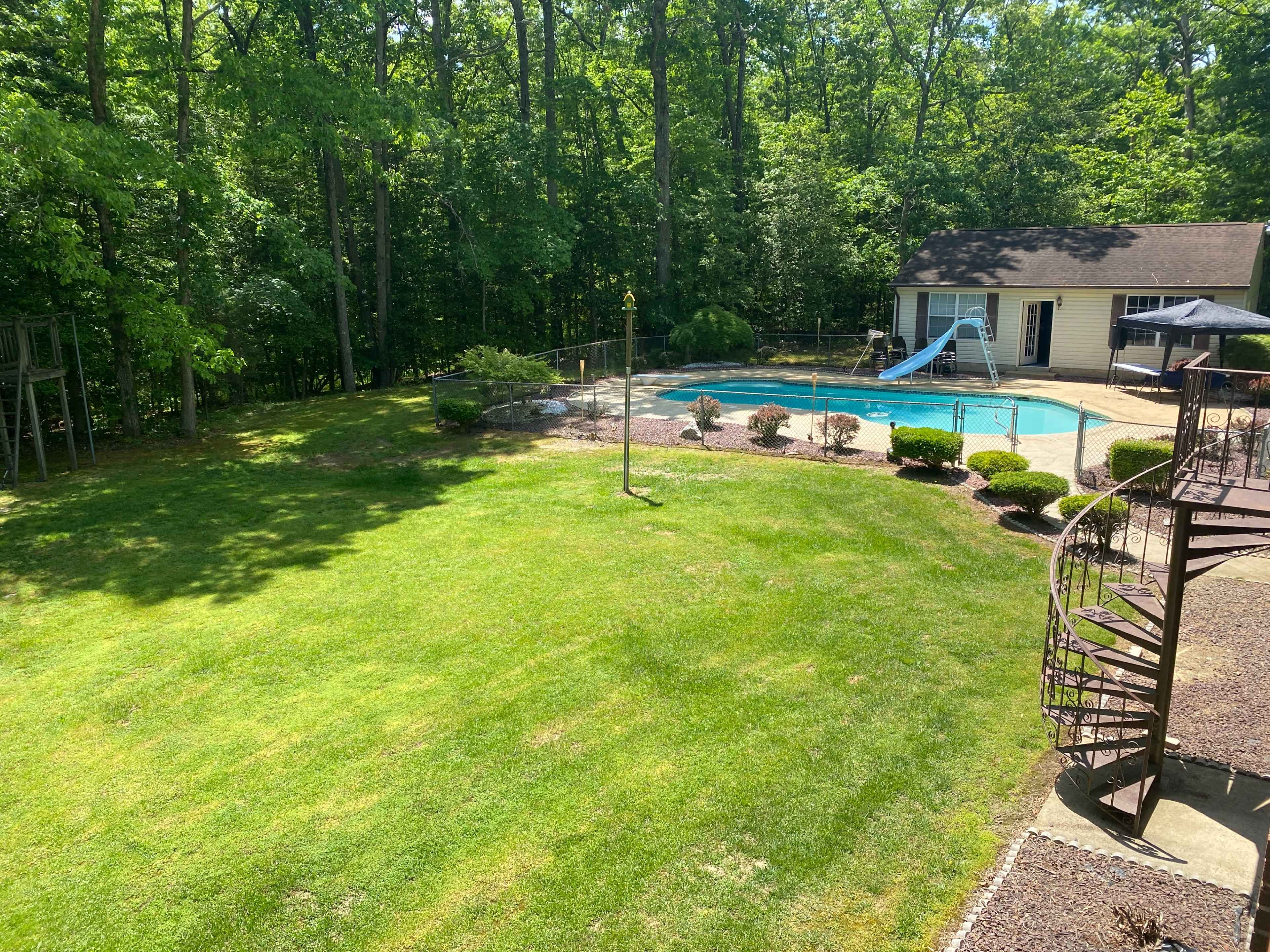A backyard with a swimming pool, a slide, landscaped bushes, and a spiral staircase leading to a deck, surrounded by trees.