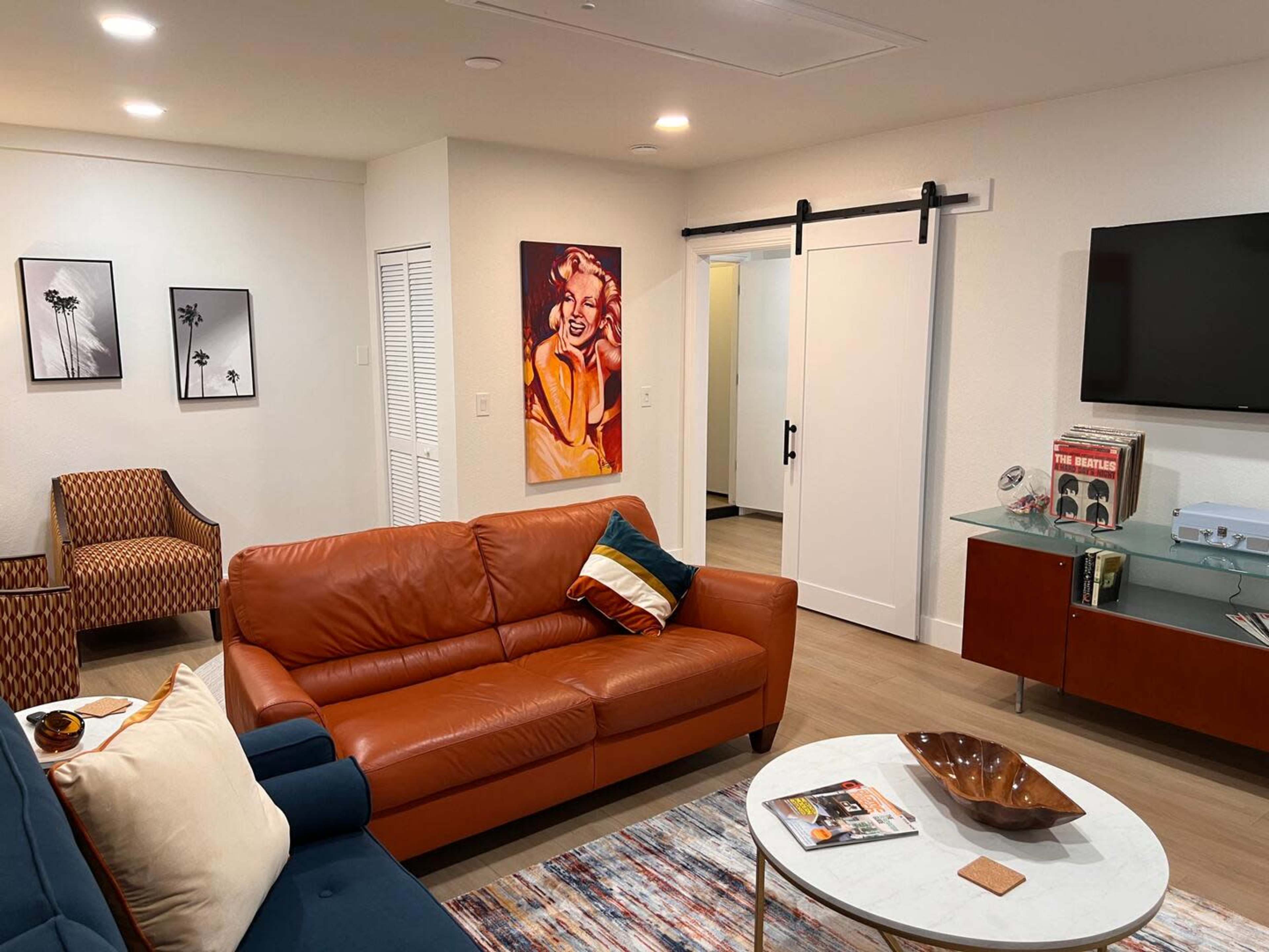 The living room features a brown leather sofa, a blue couch, a coffee table, and wall art, with a sliding door leading to another room.