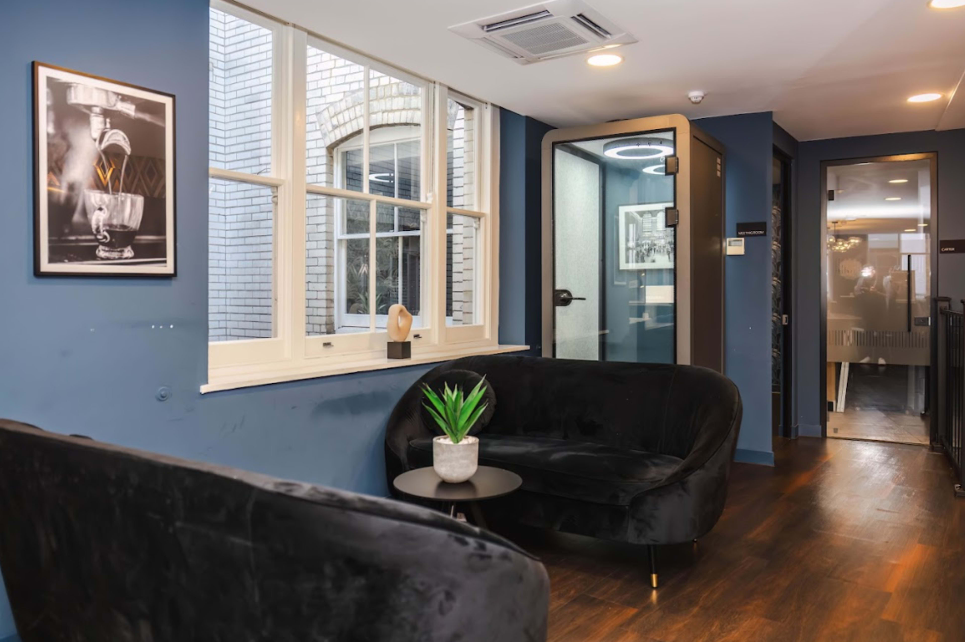 A cozy corner in an office or lounge area featuring two black velvet couches, a small round table with a potted plant, and a glass door leading to another room.