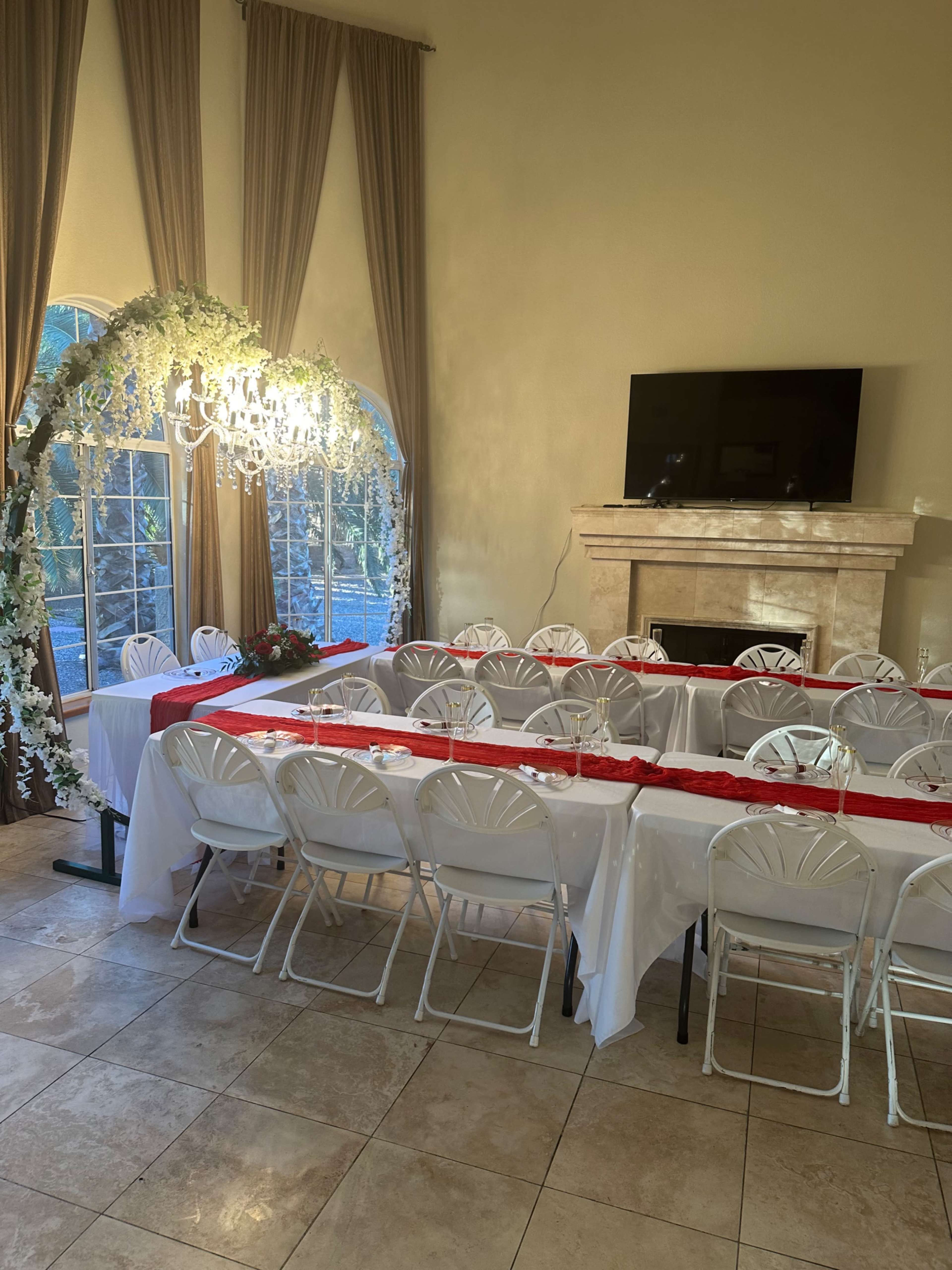 A formal dining setup with white tables and chairs is arranged under a chandelier in a room with a fireplace and large windows.