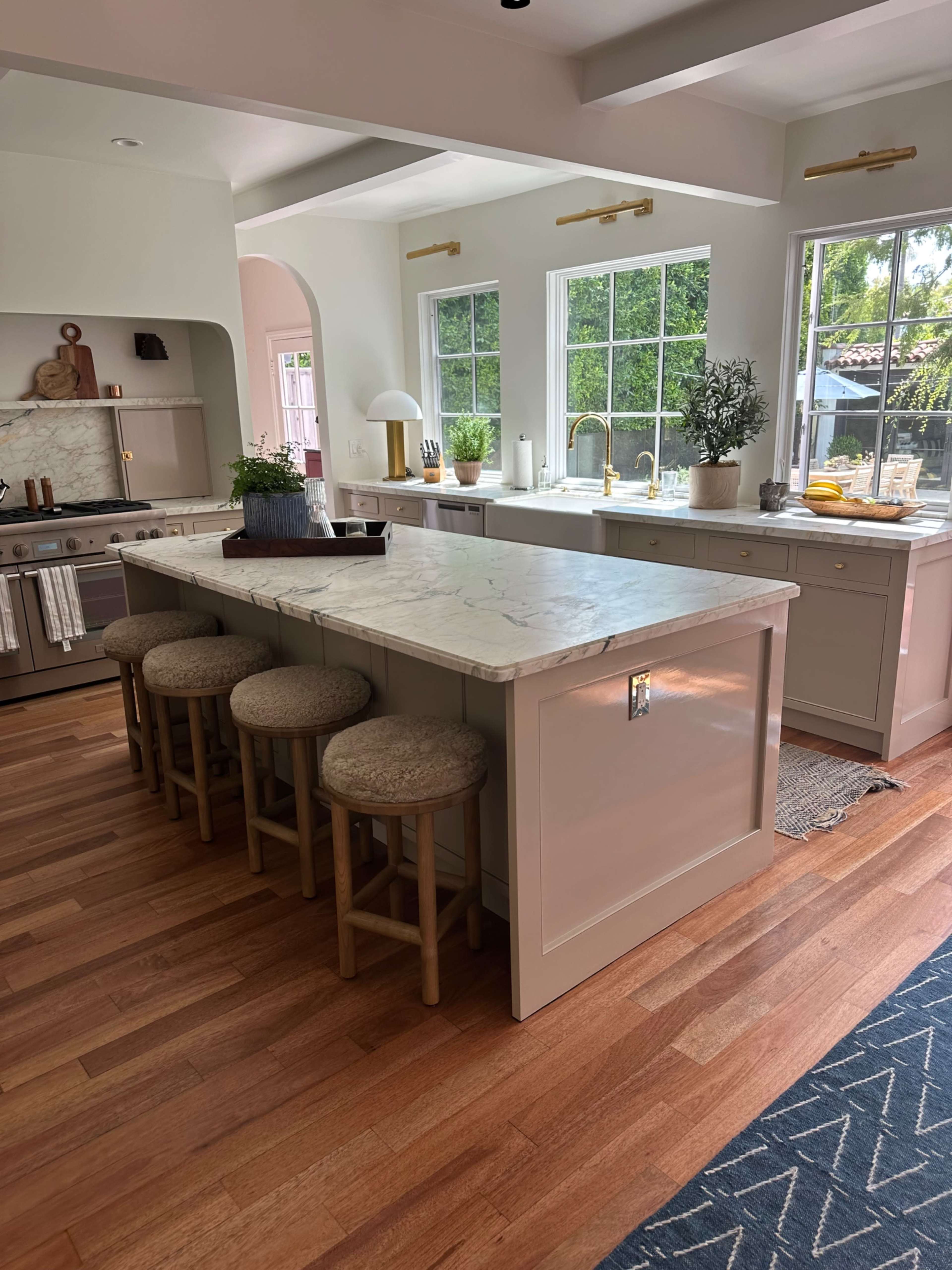 A modern kitchen features a central island with a marble countertop, four stool chairs, and large windows allowing natural light to illuminate the space.