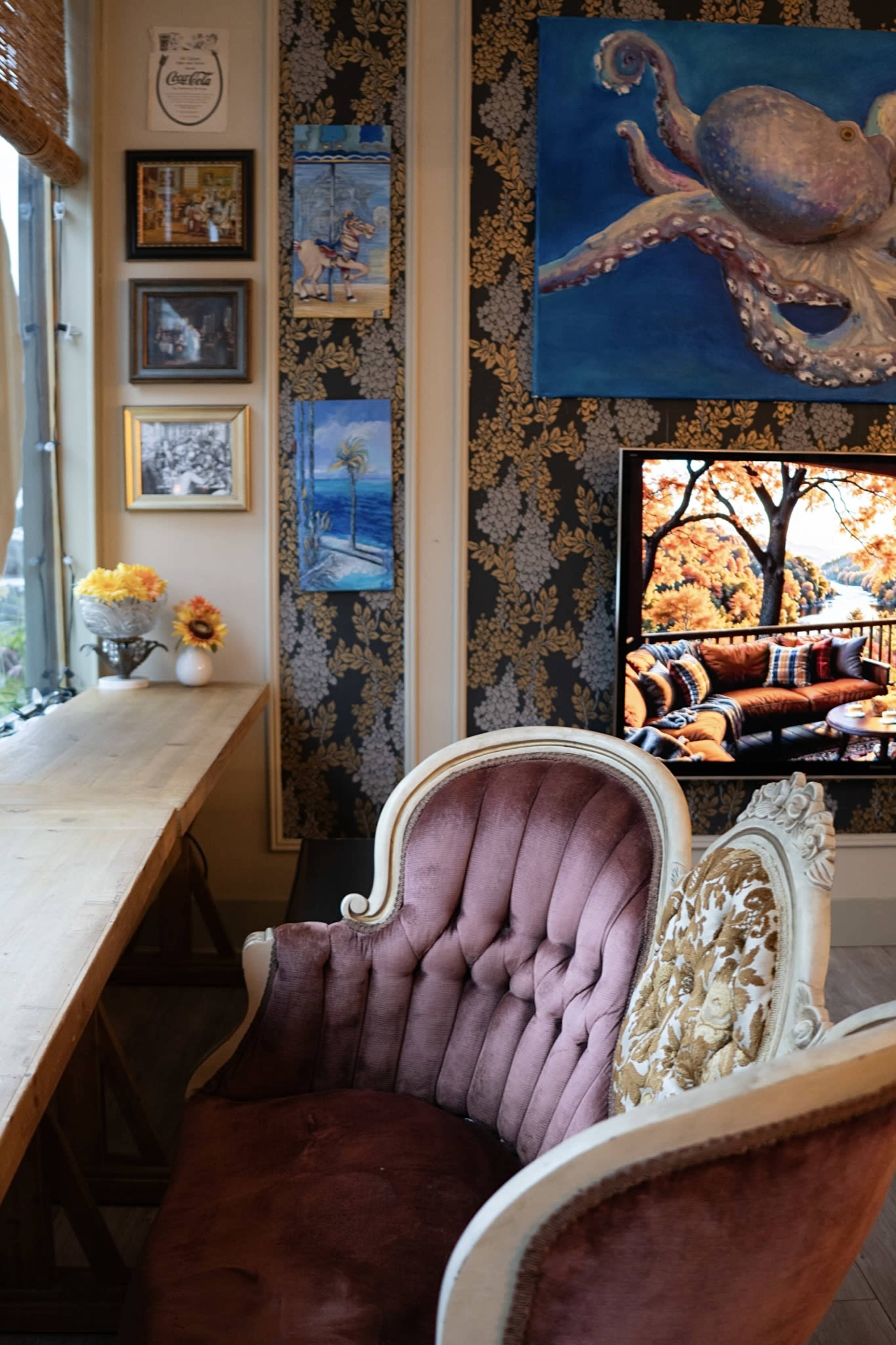 A cozy interior scene features a plush, vintage chair next to a wooden table, adorned with artwork and a colorful television displaying an autumn landscape.