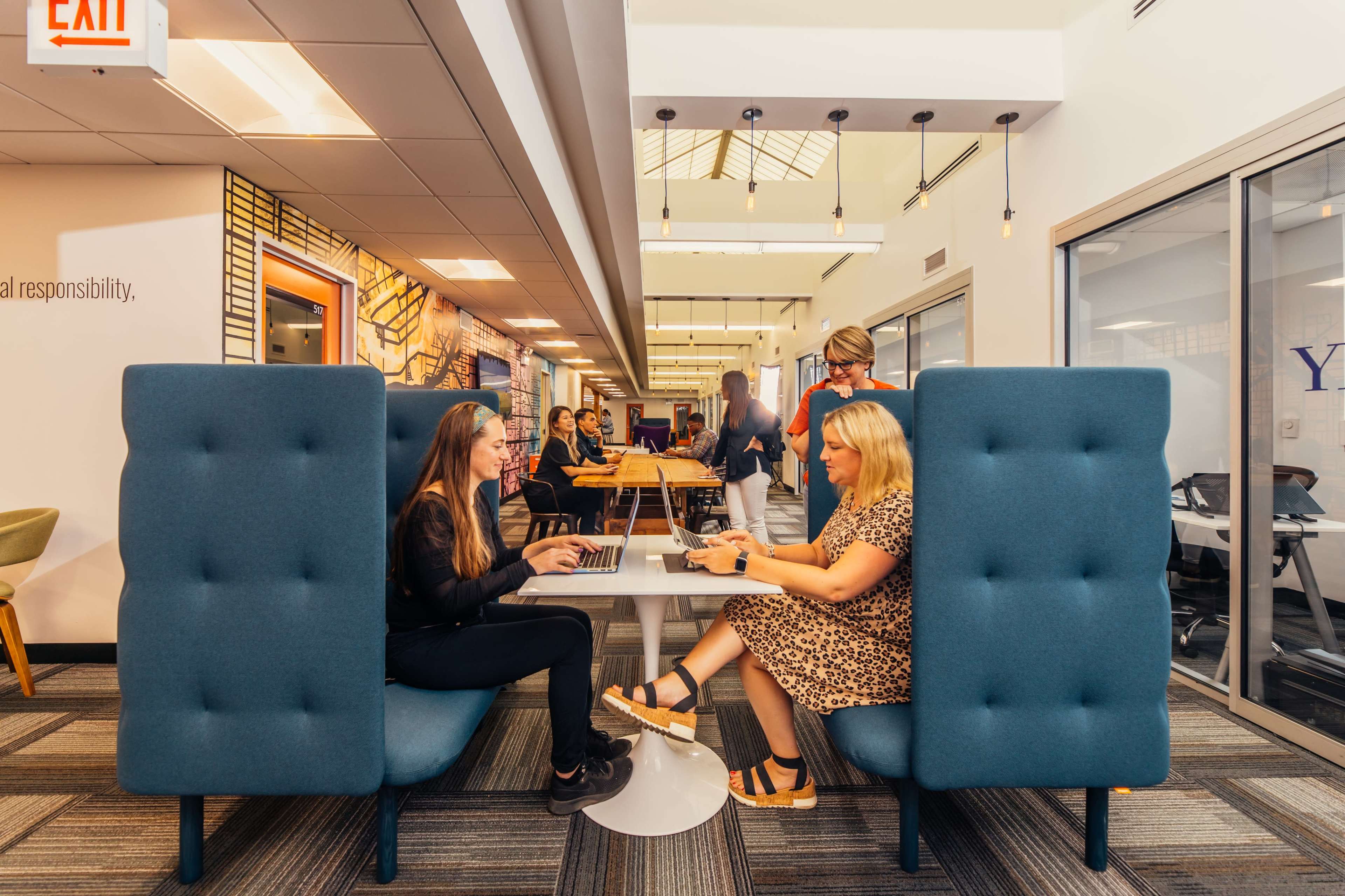 Two women are seated at a table in a modern office space, engaged in a conversation while using a laptop, with other people working in the background.