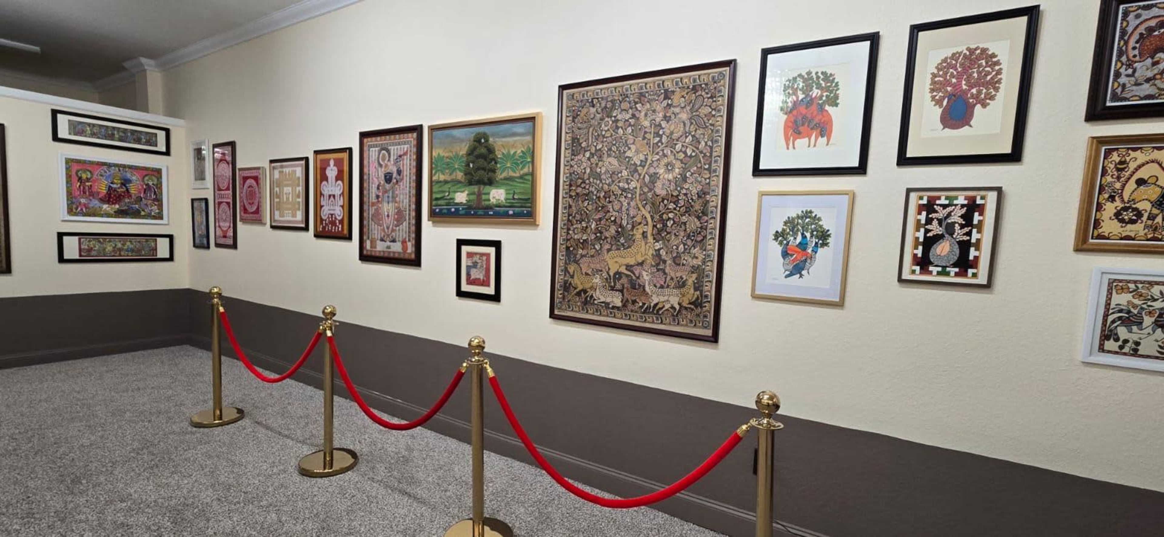 The image shows a gallery wall featuring various framed artworks and decorative pieces, separated by red rope barriers.