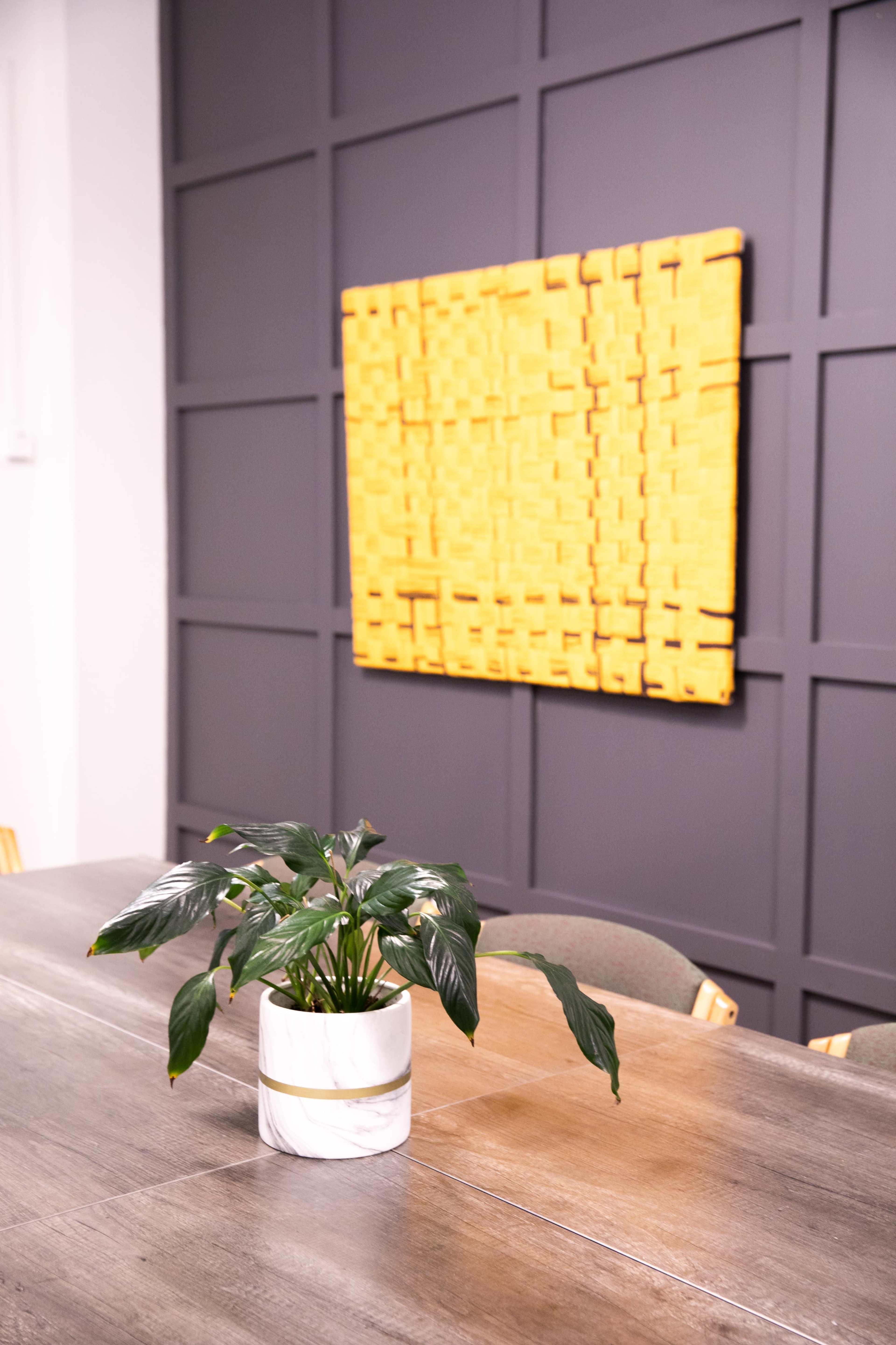 A small potted plant sits on a wooden table in front of a textured yellow wall art piece.