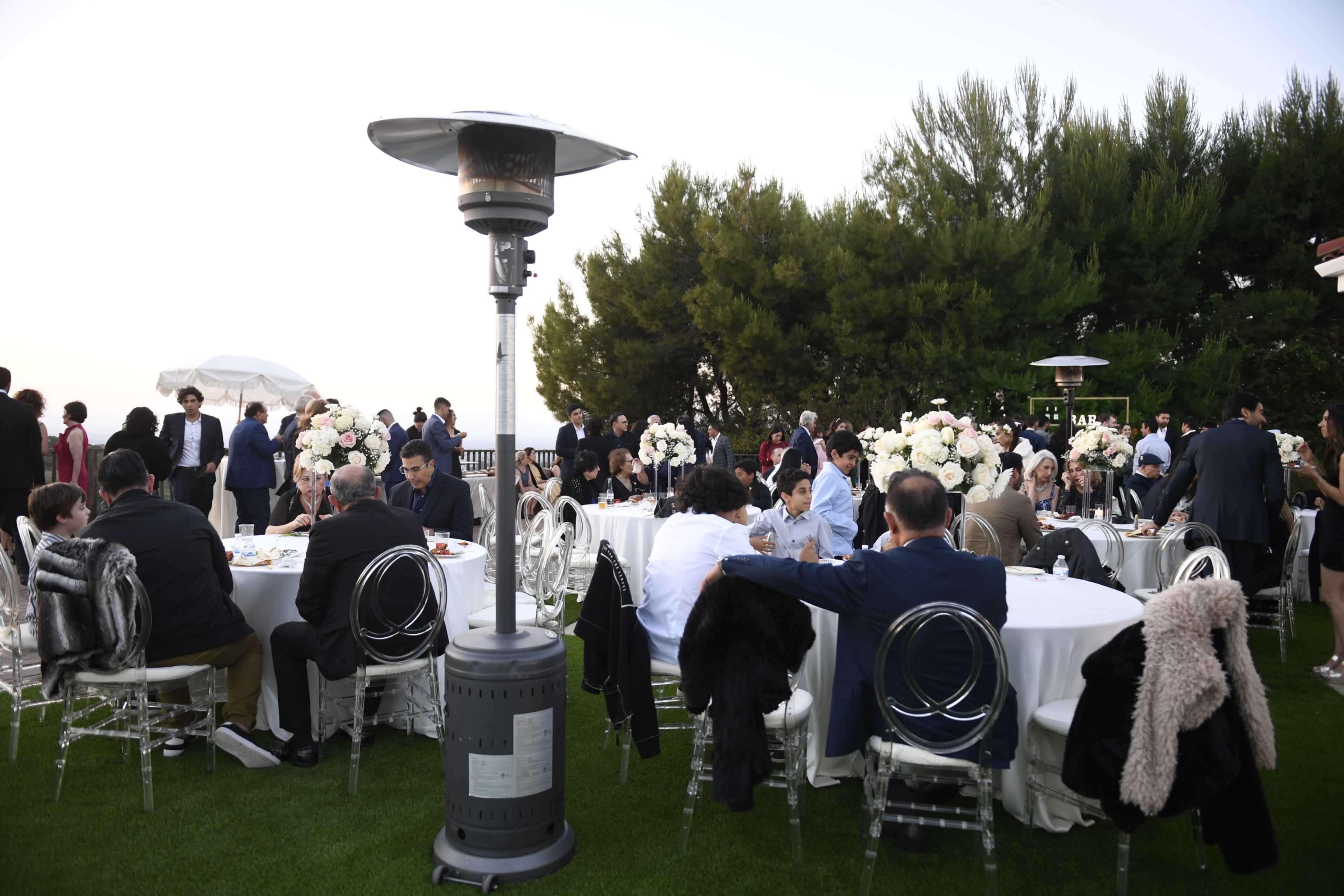 People are gathered around elegantly set tables at an outdoor event surrounded by greenery.