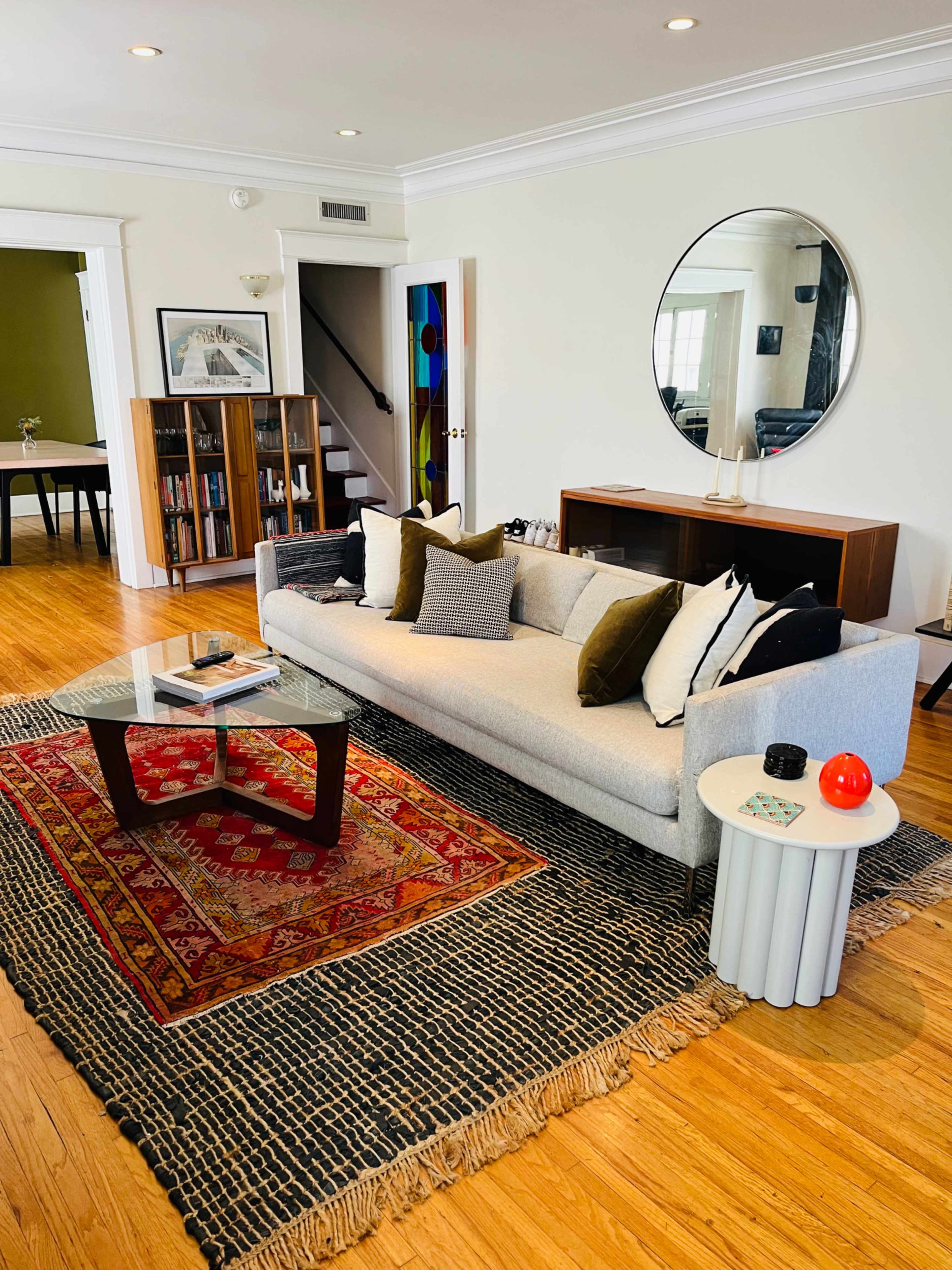 The living room features a gray sofa with several decorative pillows, a glass coffee table on a patterned rug, and a console table against the wall with bookshelves in the background.