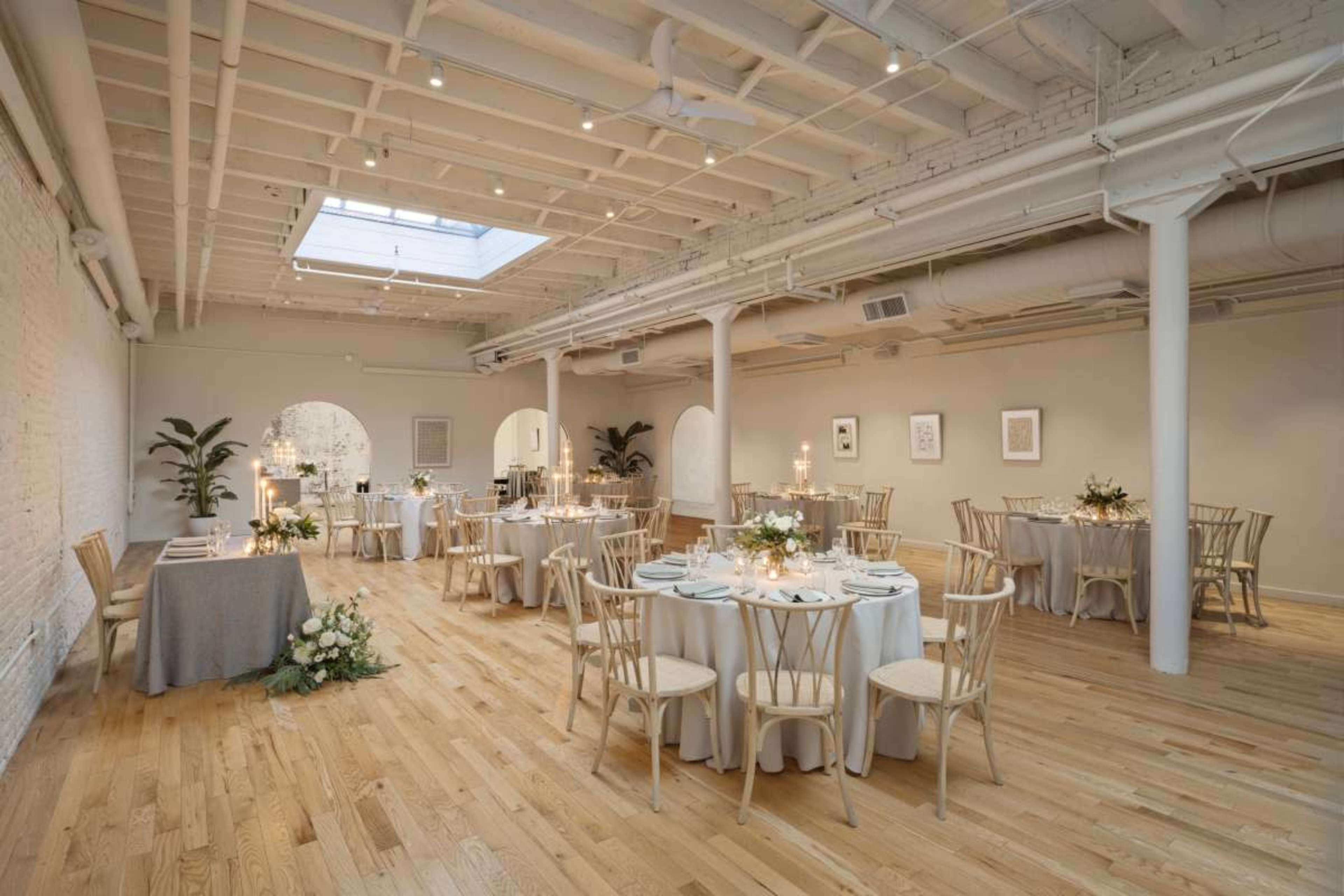 The image shows a beautifully arranged event space with round tables set for dining, soft lighting, and plants in each corner.