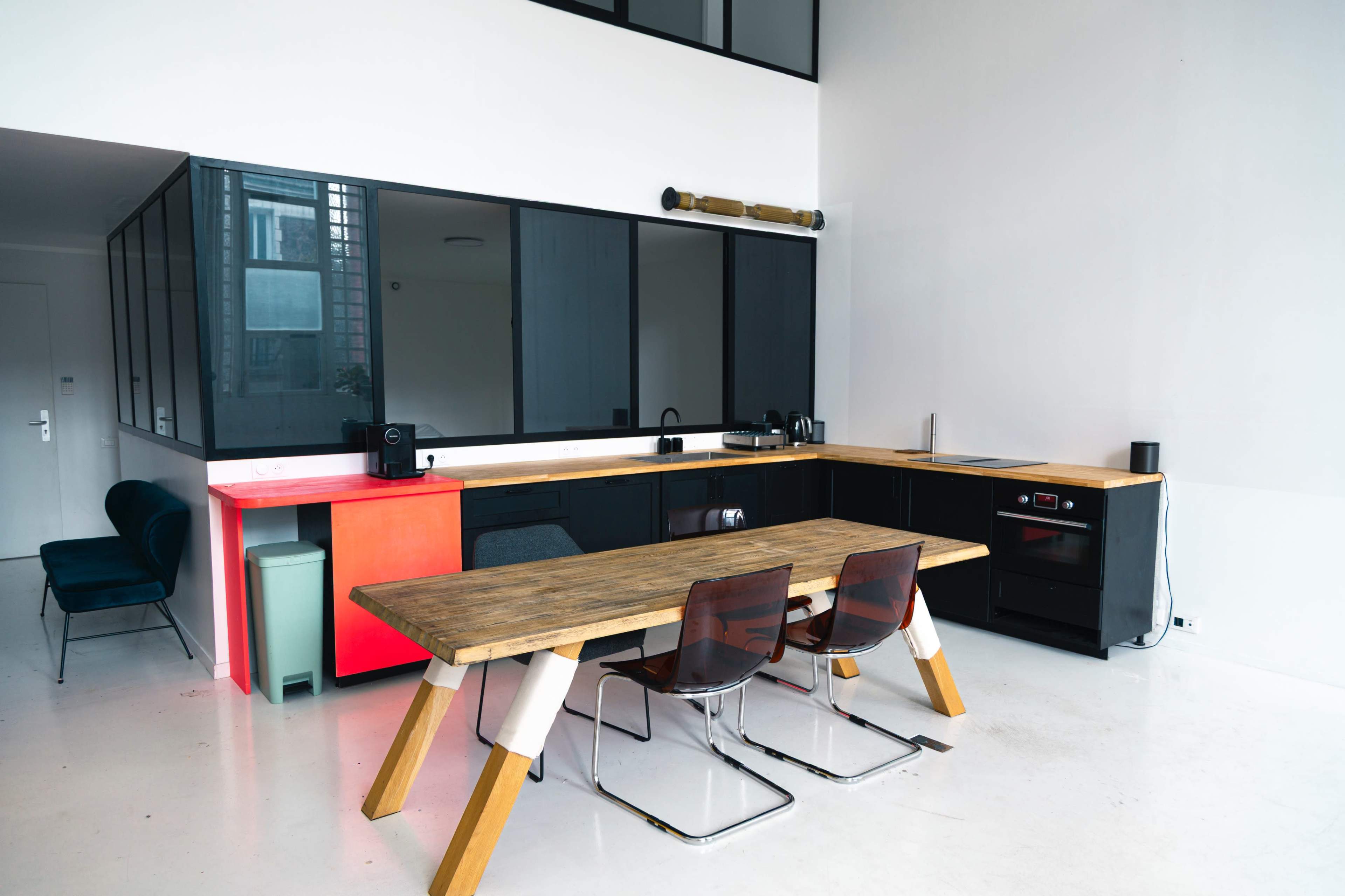 The image shows a modern kitchen with a large wooden table and transparent chairs, set against a backdrop of sleek cabinets and large windows.