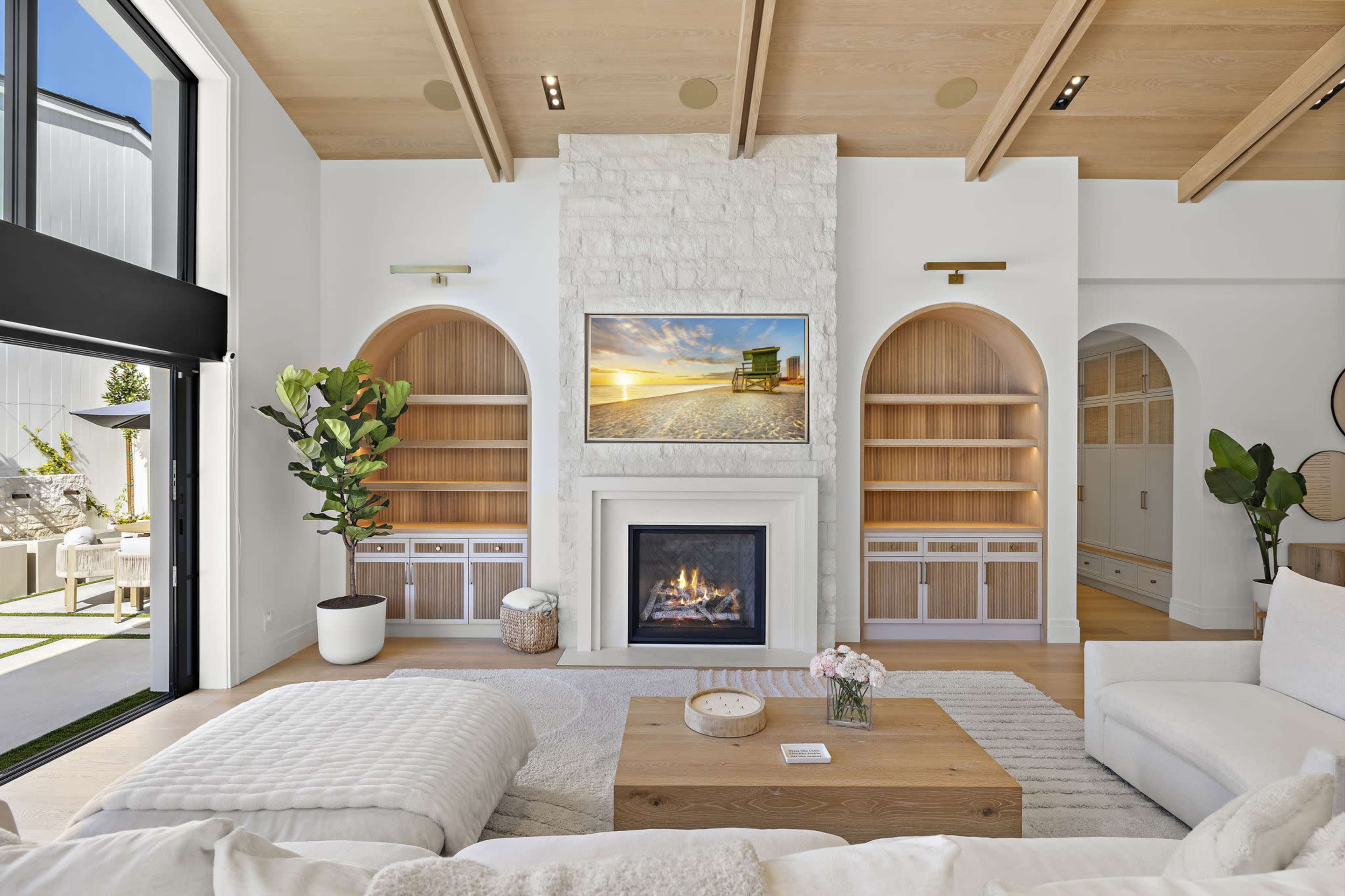 The image shows a modern living room with a stone fireplace, built-in shelves, and a large television above the mantel, illuminated by natural light.