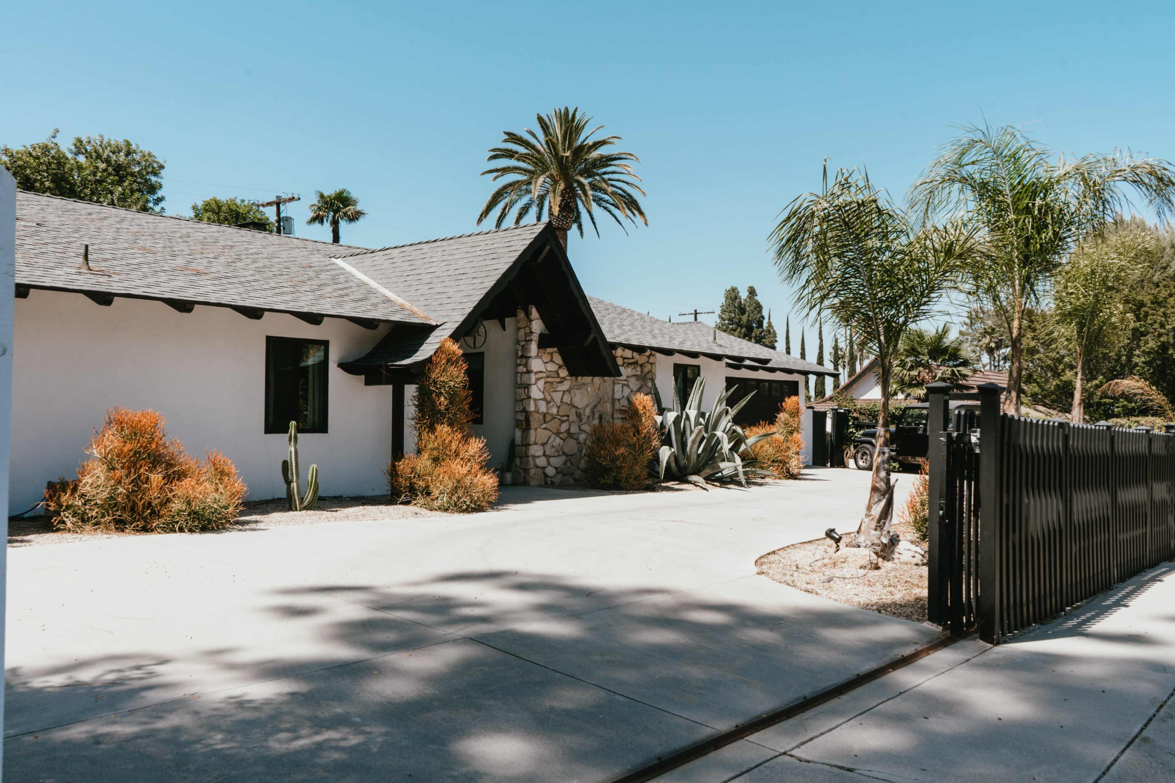 The image shows a single-story house with a stone facade, surrounded by palm trees and shrubs, set against a clear blue sky.