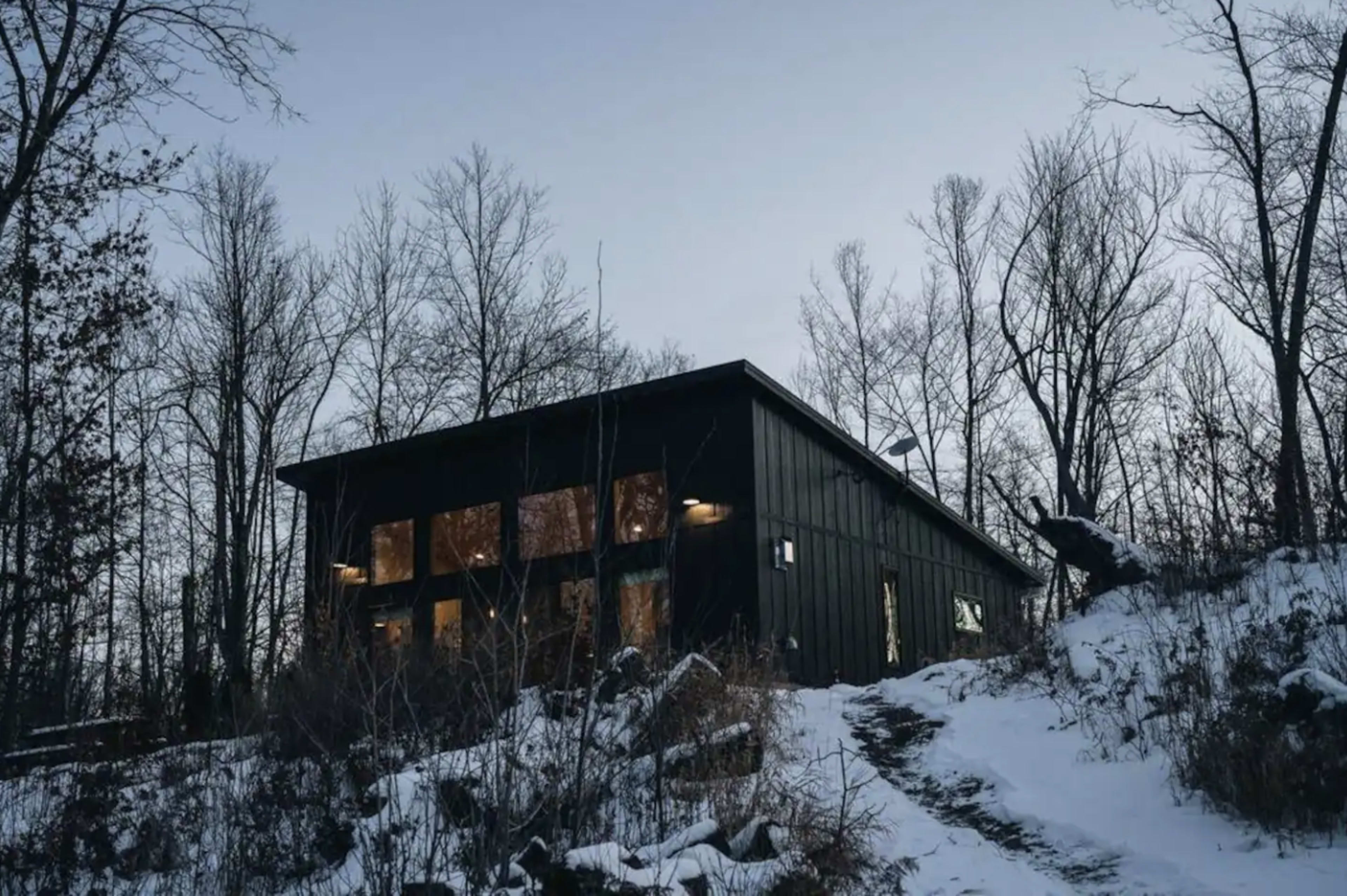A black modern house sits on a snow-covered slope, surrounded by bare trees against a twilight sky.