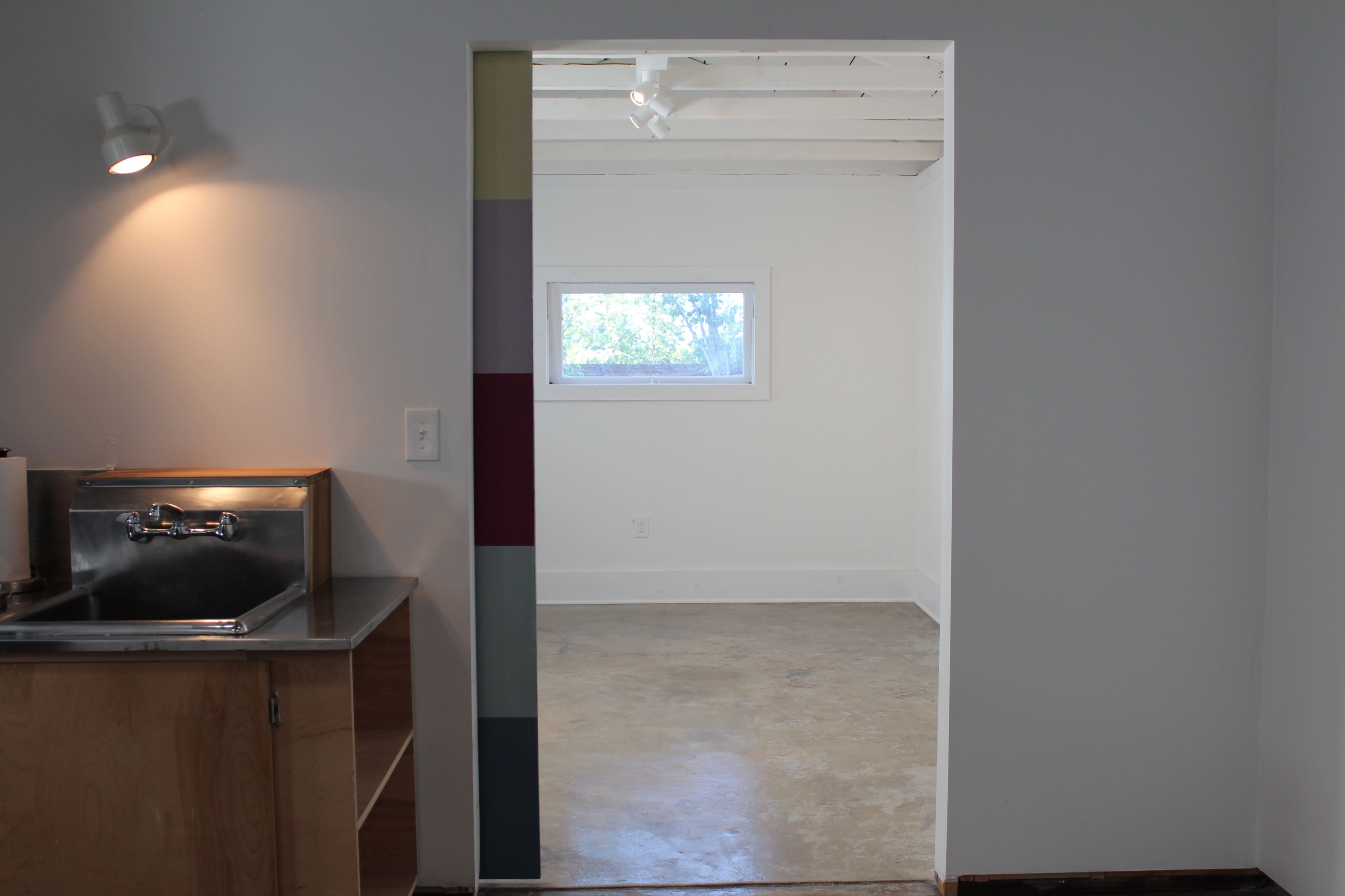 The image shows a small, minimalist room with a kitchenette to the left and an empty space beyond a doorway on the right.