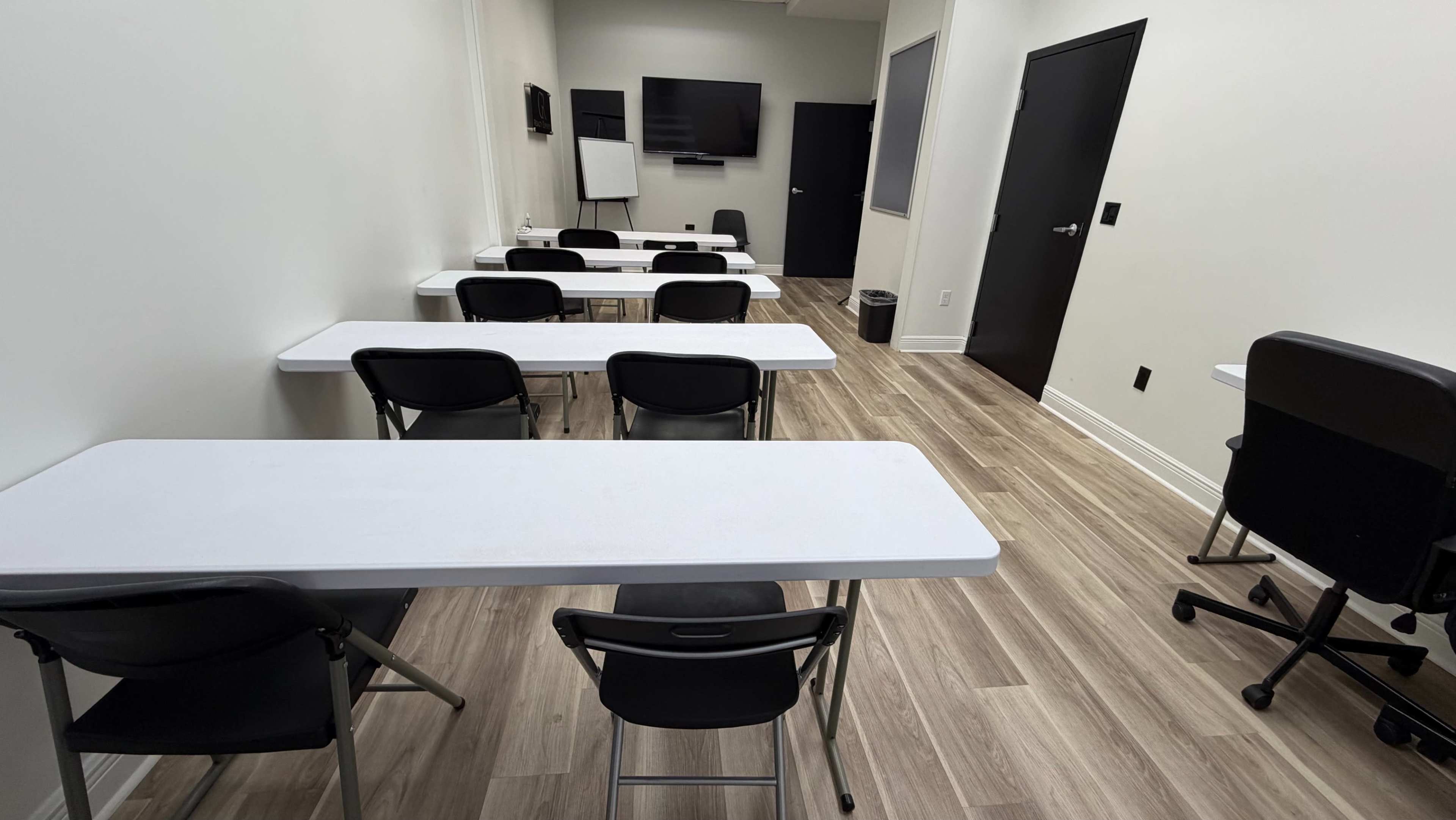 The image shows a classroom with several white tables and black chairs arranged in rows, along with a television and whiteboard mounted on the wall.