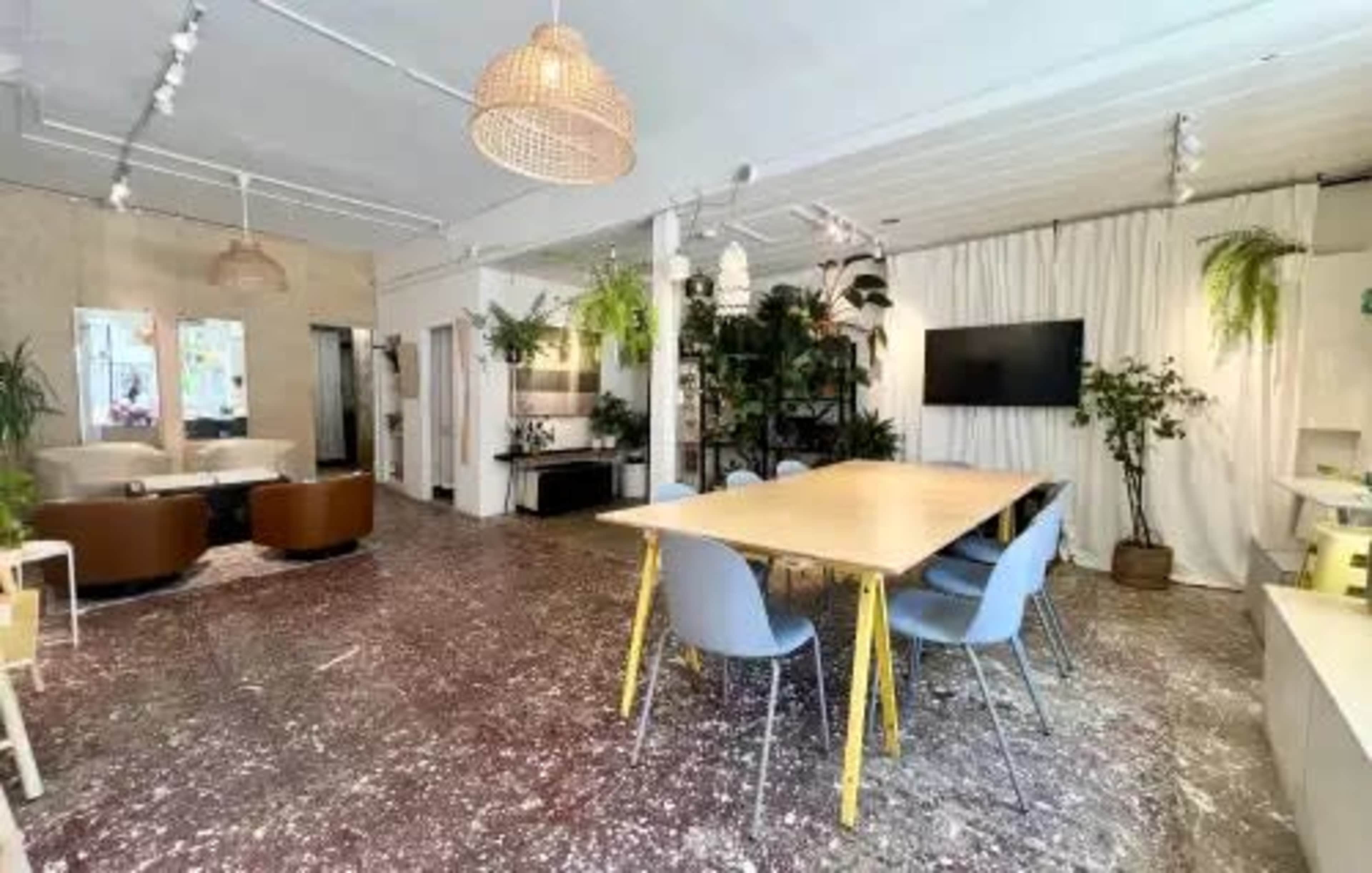 The image shows a modern office space with a large wooden table surrounded by chairs, plants, and a small seating area, illuminated by natural light.