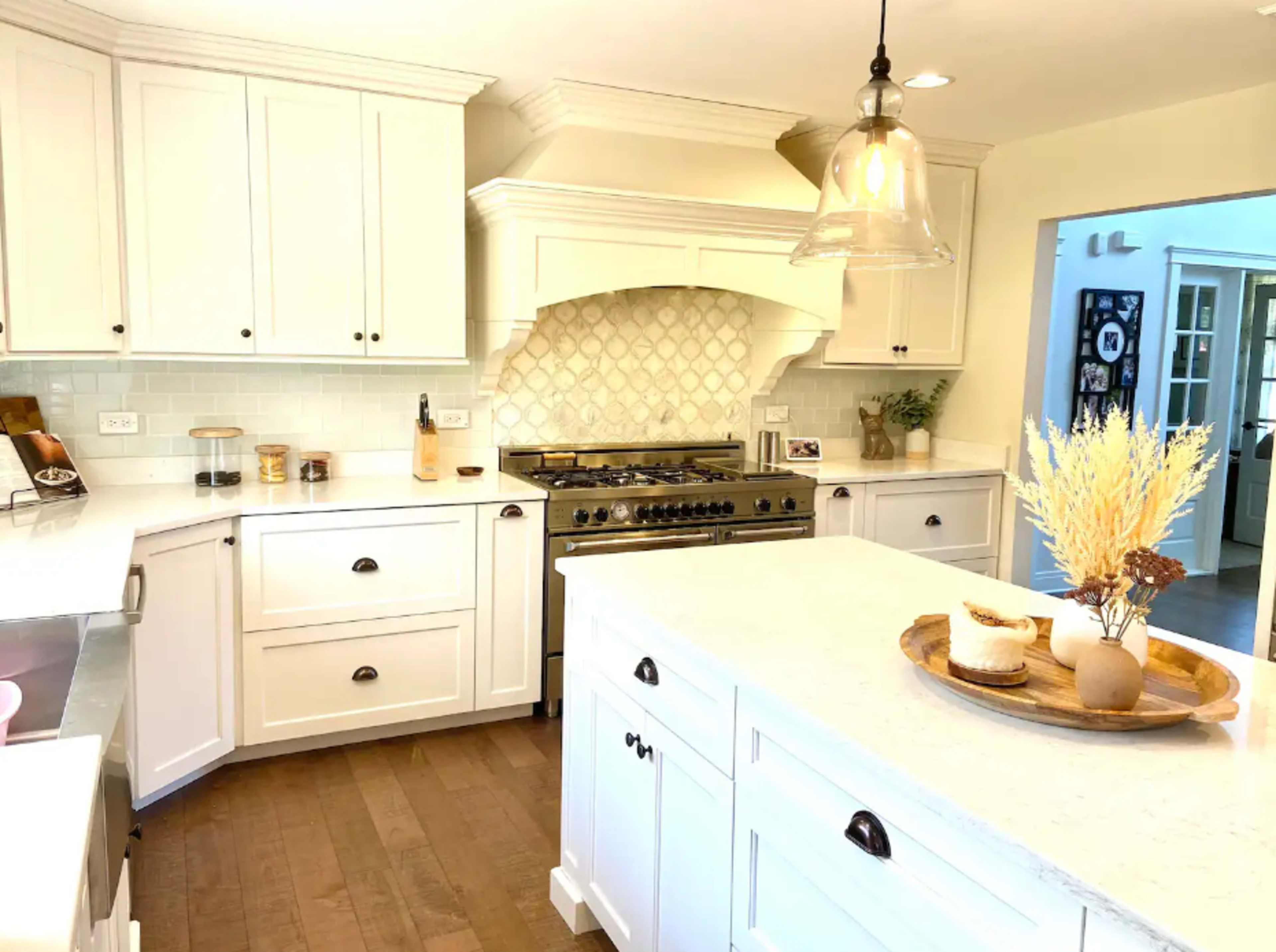 A modern kitchen features white cabinetry, a large island with a wooden tray, and a decorative range hood above a stainless steel stove.