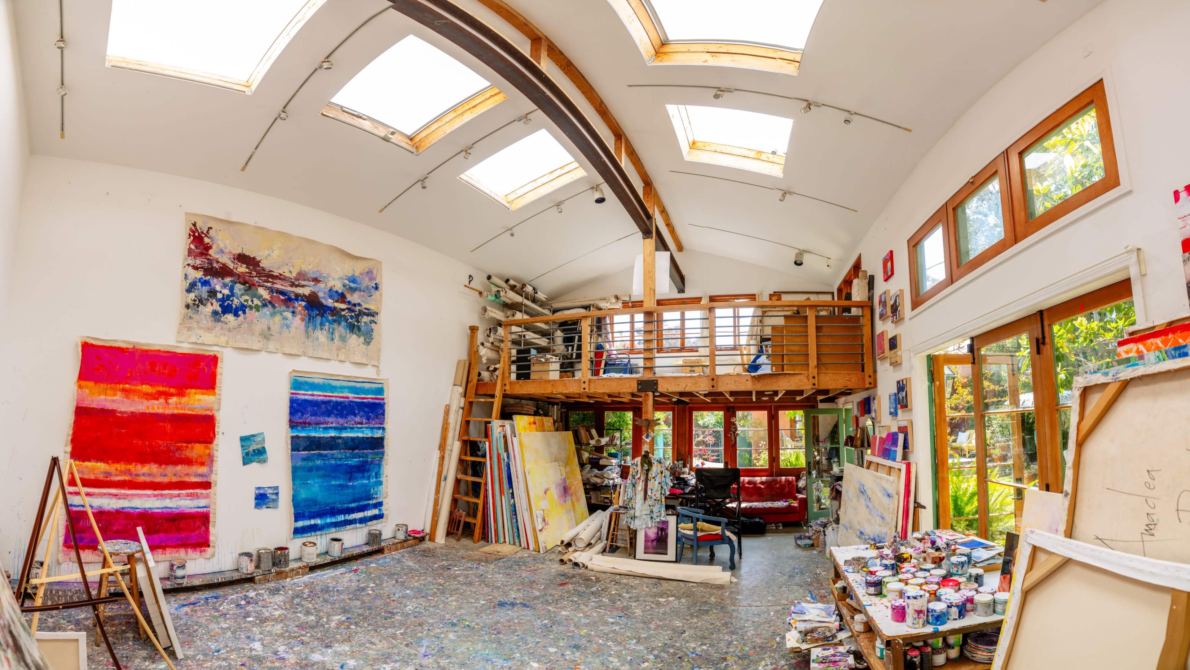 A spacious art studio with large skylights, featuring colorful paintings on the walls and various art supplies scattered on the floor.