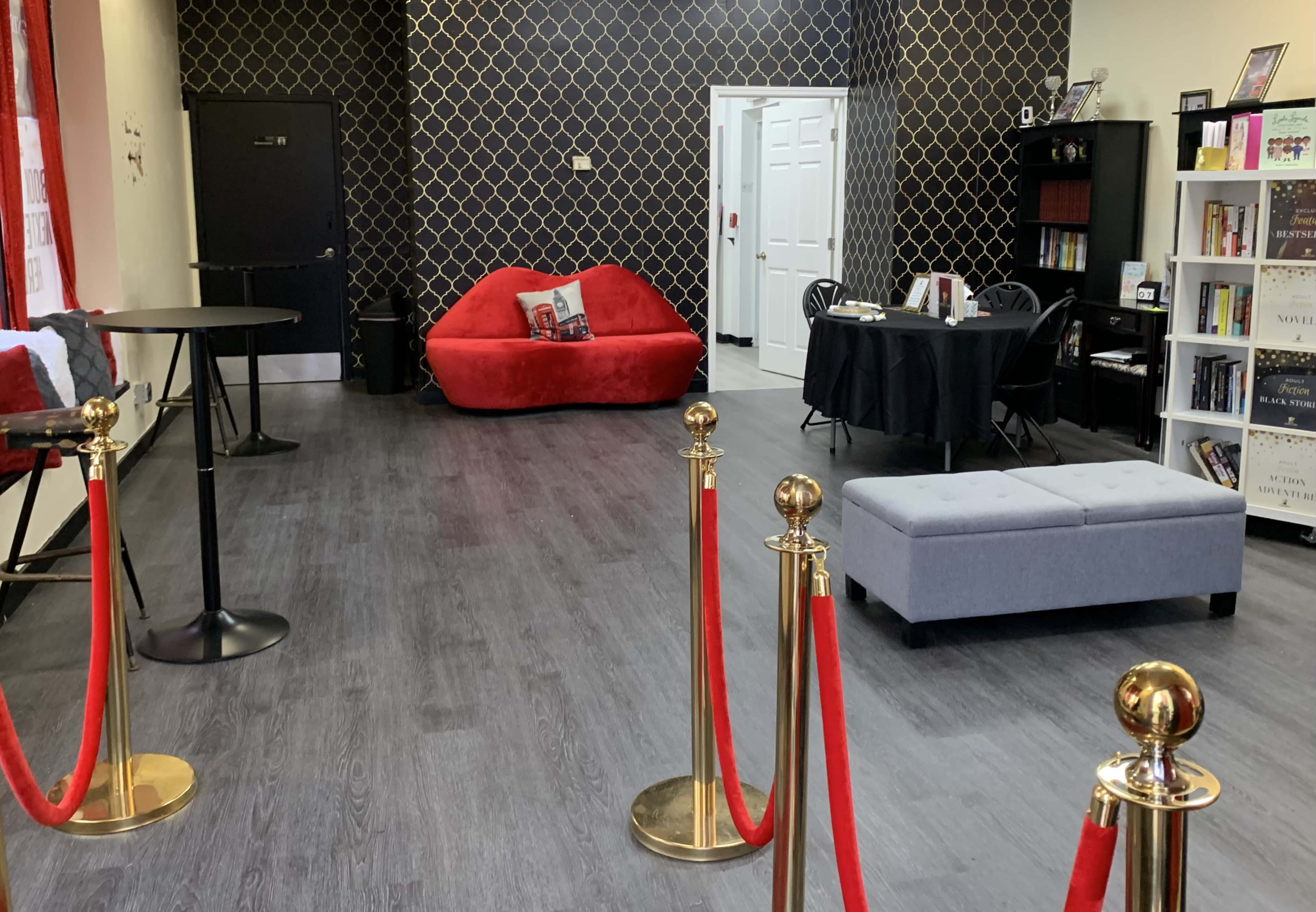 The image shows a modern interior space featuring a red couch, a grey ottoman, a black table and chairs, and a bookshelf, with red rope barriers in the foreground.