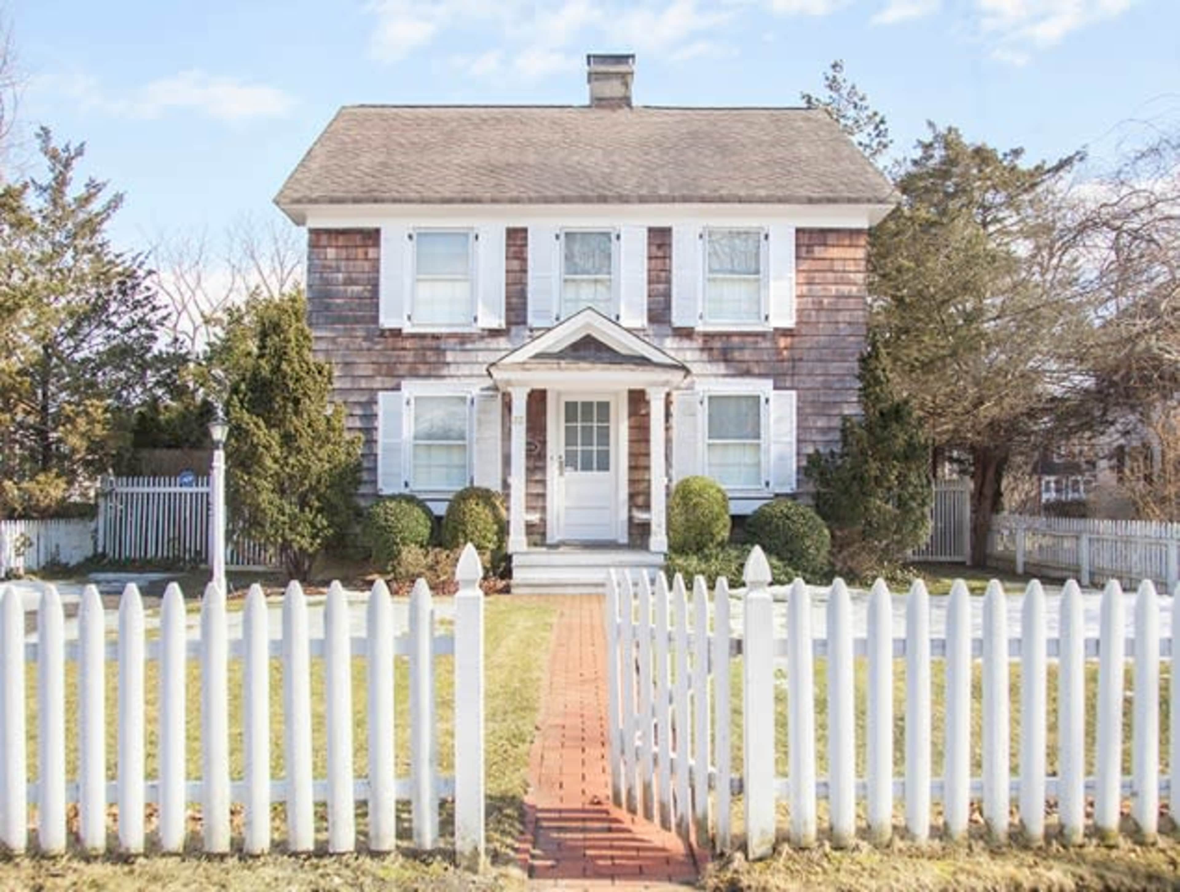 A two-story house with a shingled exterior, white shutters, and a picket fence surrounds a manicured front yard.