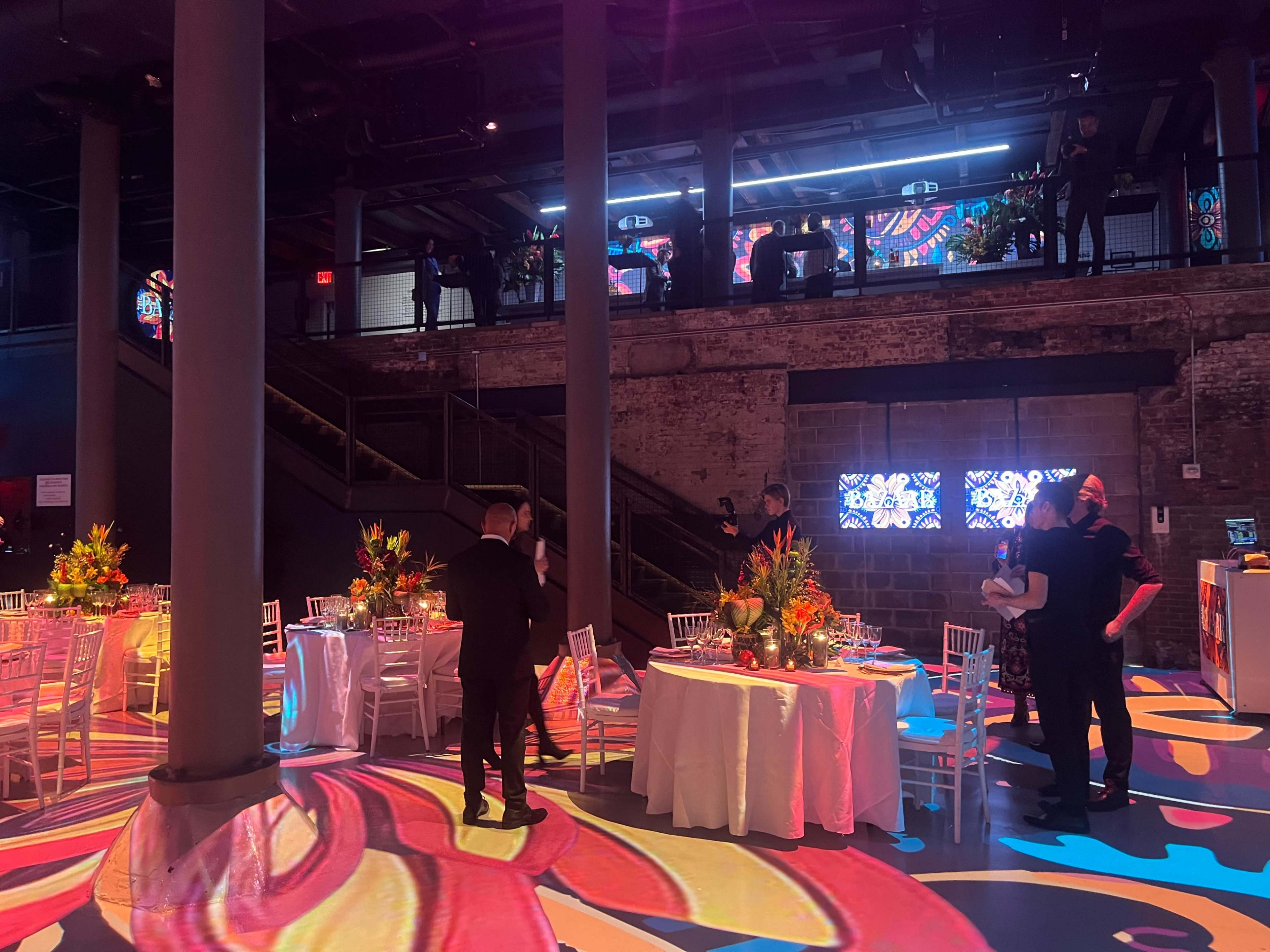 A vibrant event space features decorated tables and guests interacting, with colorful projections illuminating the floor and walls.