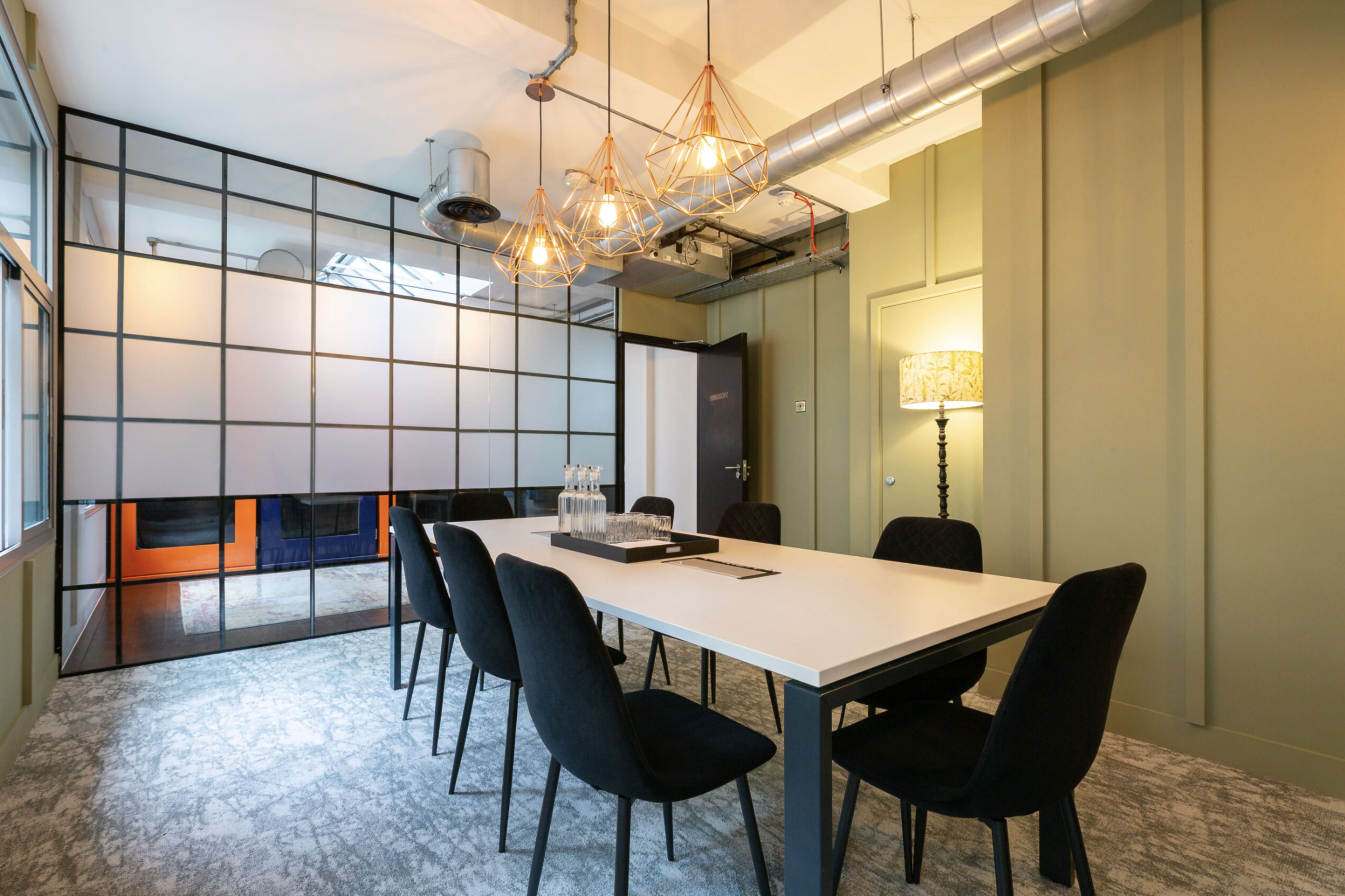 The image shows a modern meeting room featuring a long white table surrounded by black chairs, with large pendant lights overhead and glass partitions creating a sophisticated atmosphere.