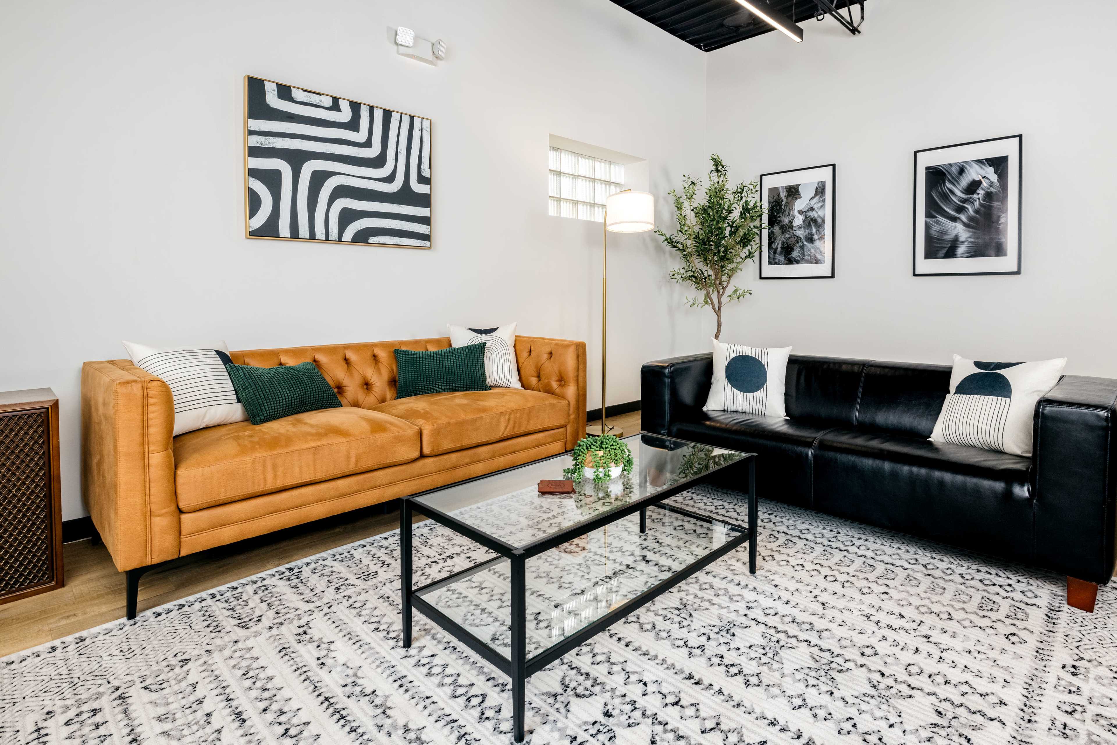 A modern living room with a black leather sofa, a tan upholstered couch, a glass coffee table, and framed artwork on the walls.