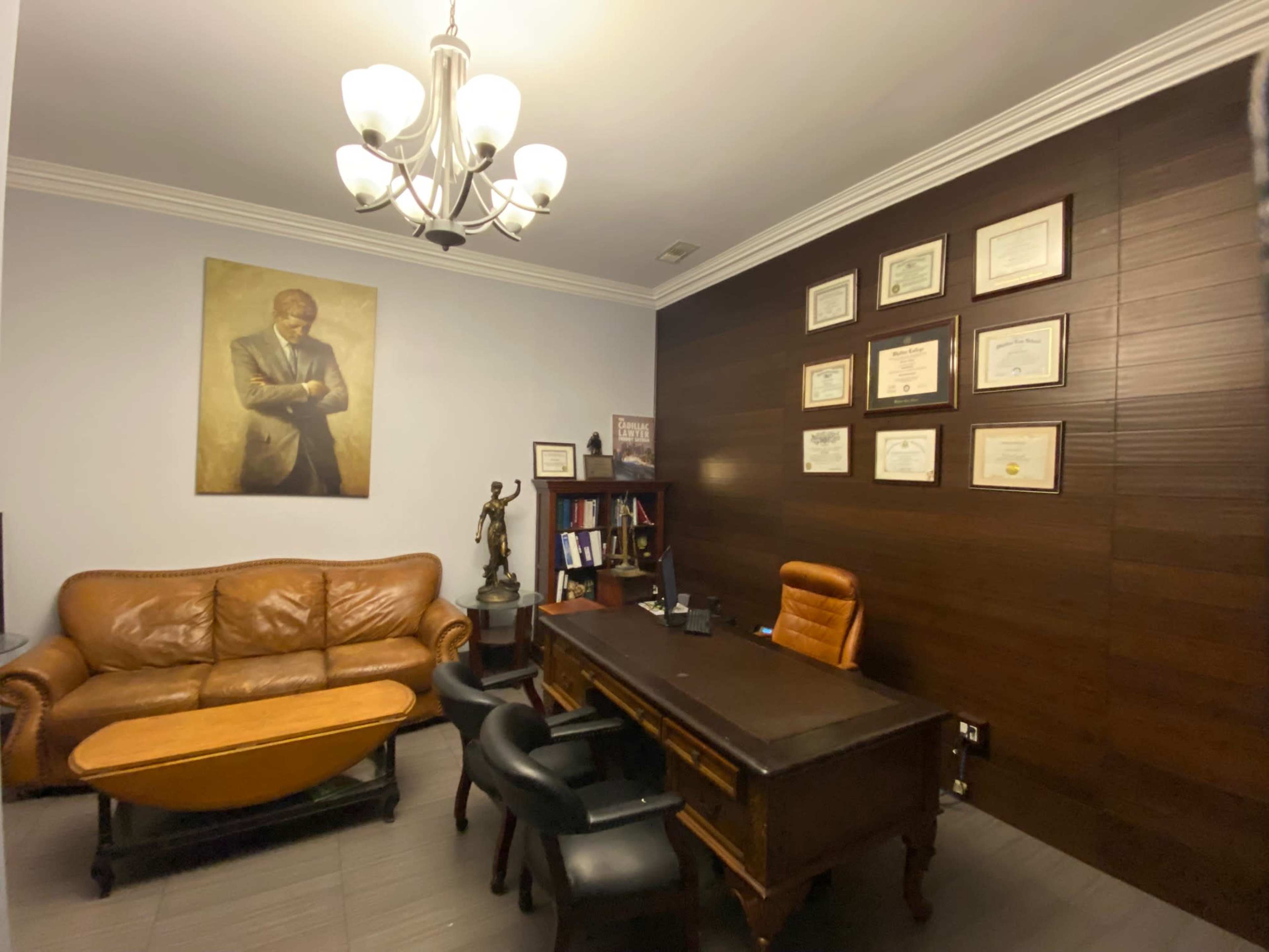 A well-furnished office with a brown sofa, a wooden desk, bookshelves filled with books and framed certificates on the wall.