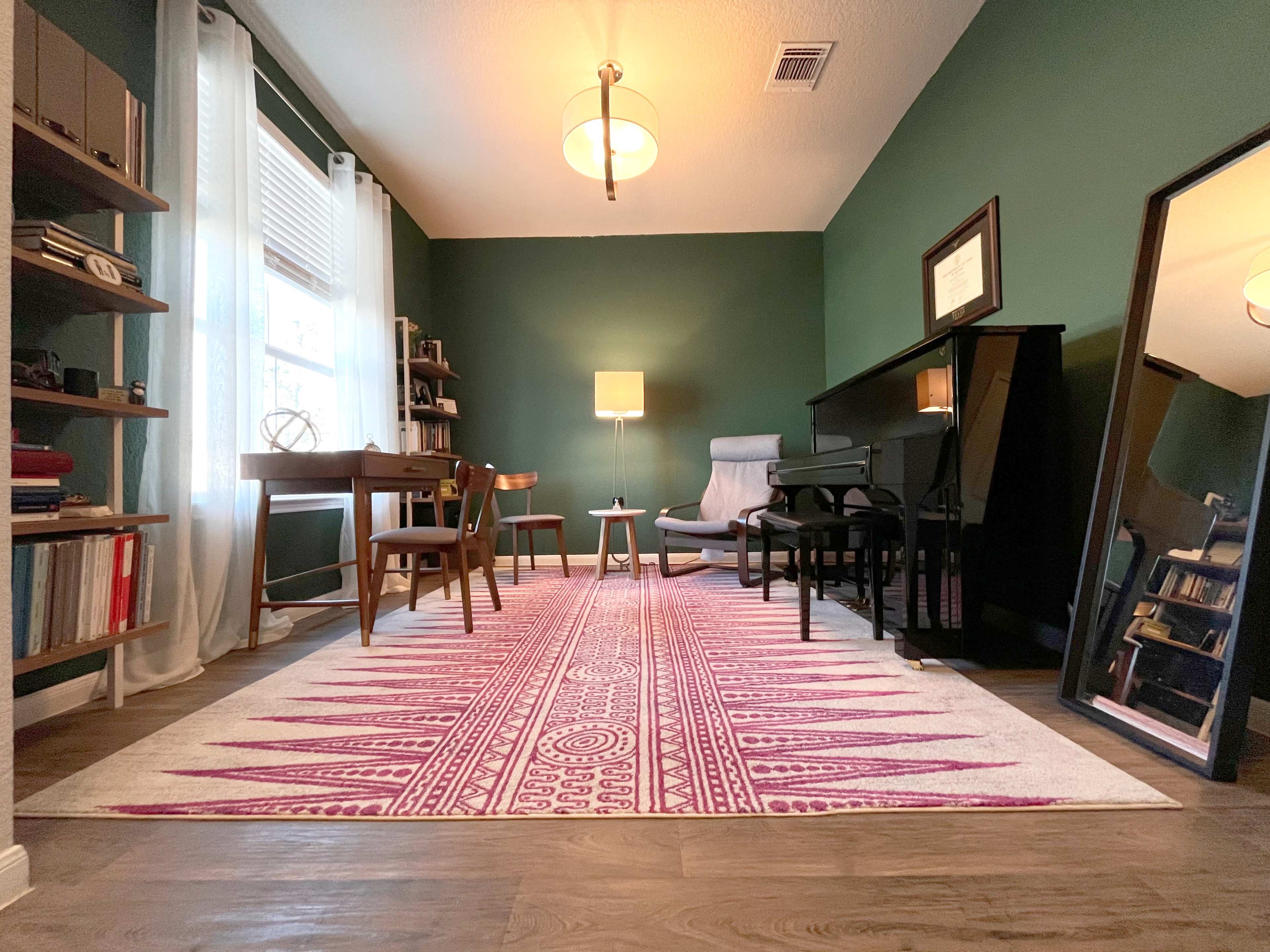 The scene depicts a cozy room with a green wall, featuring a desk, chairs, a piano, a lamp, and a large mirror, all arranged over a patterned rug.