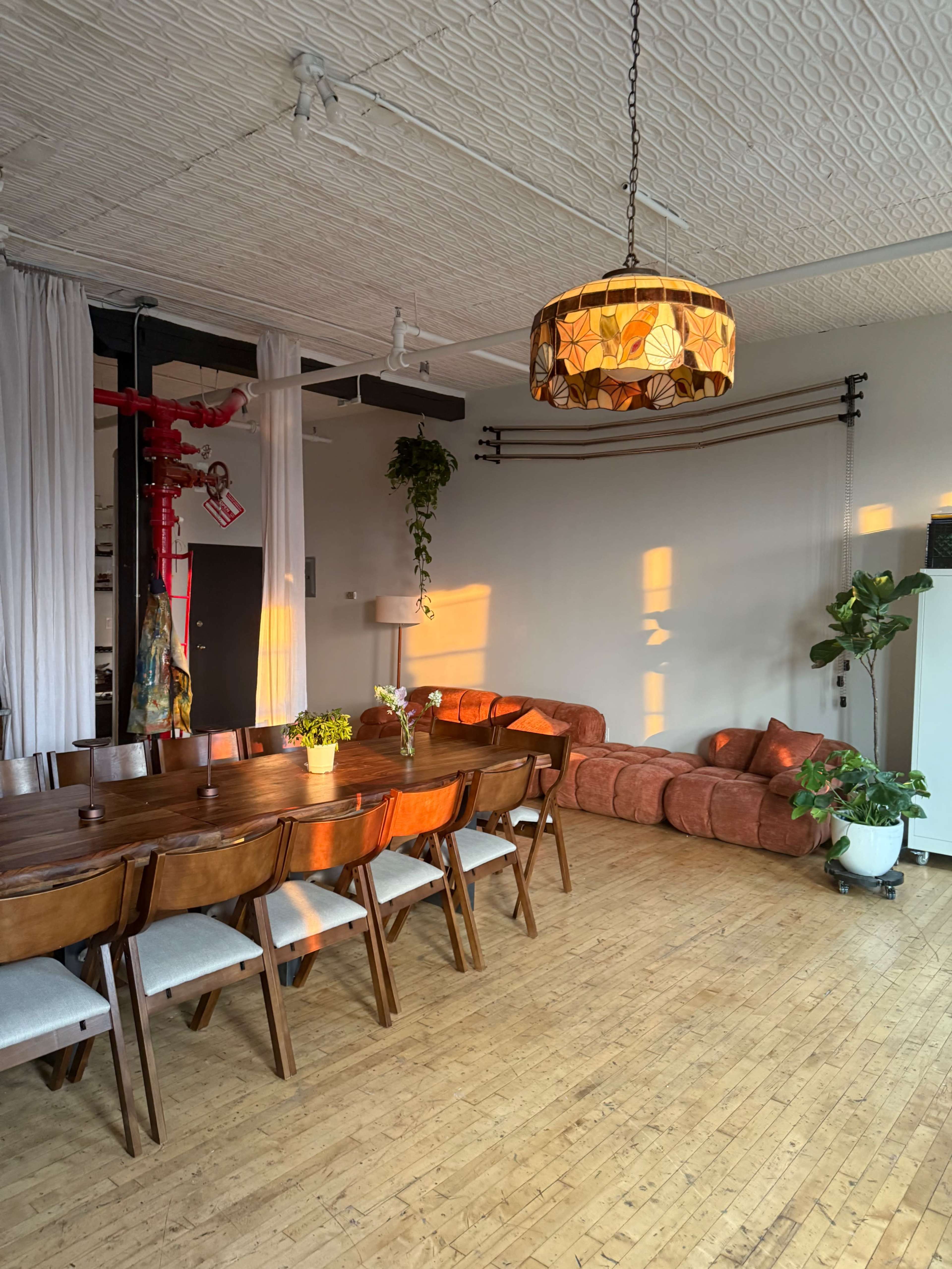 A large dining table with wooden chairs is positioned next to a cozy seating area and illuminated by warm light from a lamp, in a modern, spacious room featuring wooden floors.