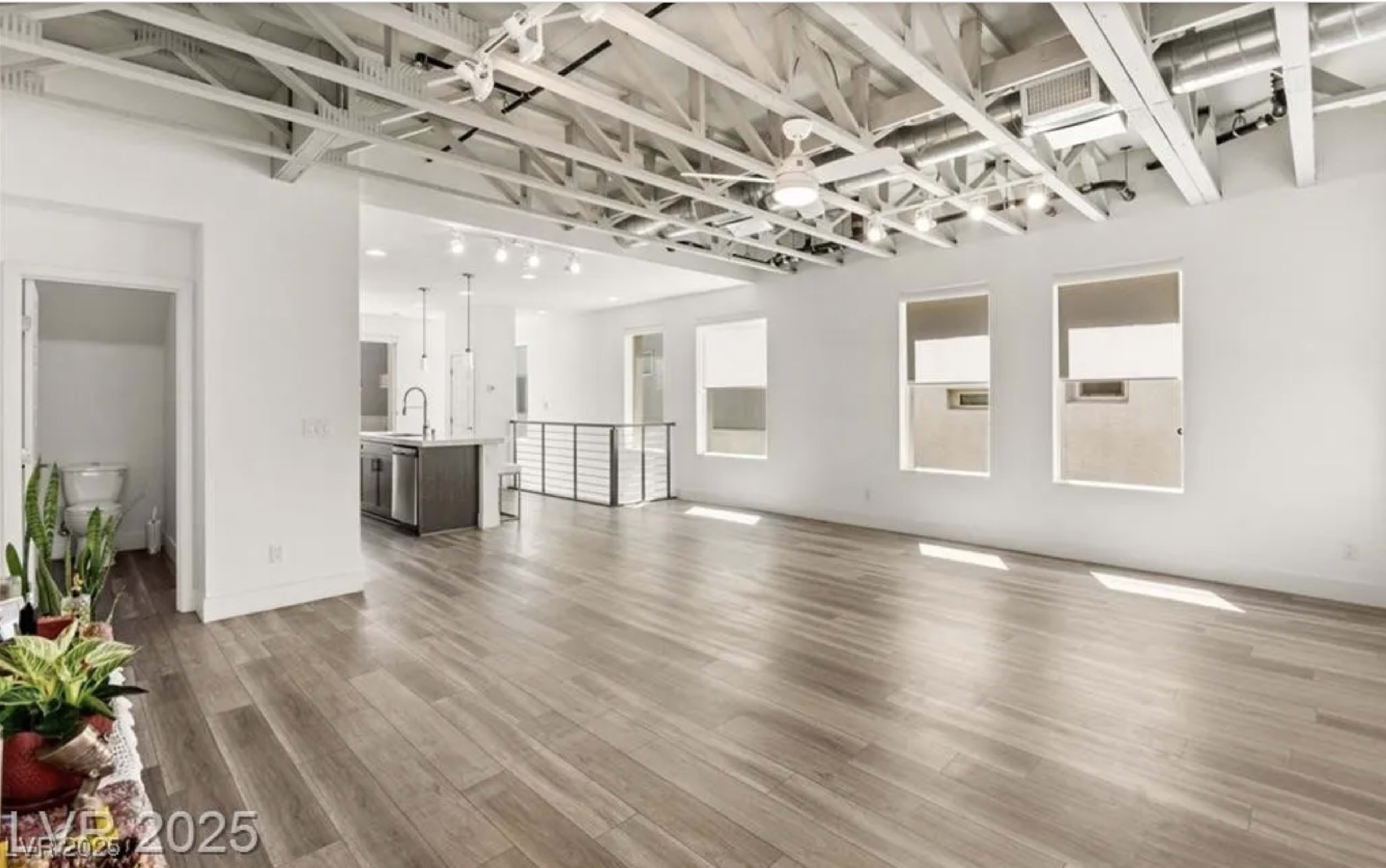 The image shows an open-concept living space with a modern kitchen, featuring wooden floors, exposed beams, and large windows allowing natural light.