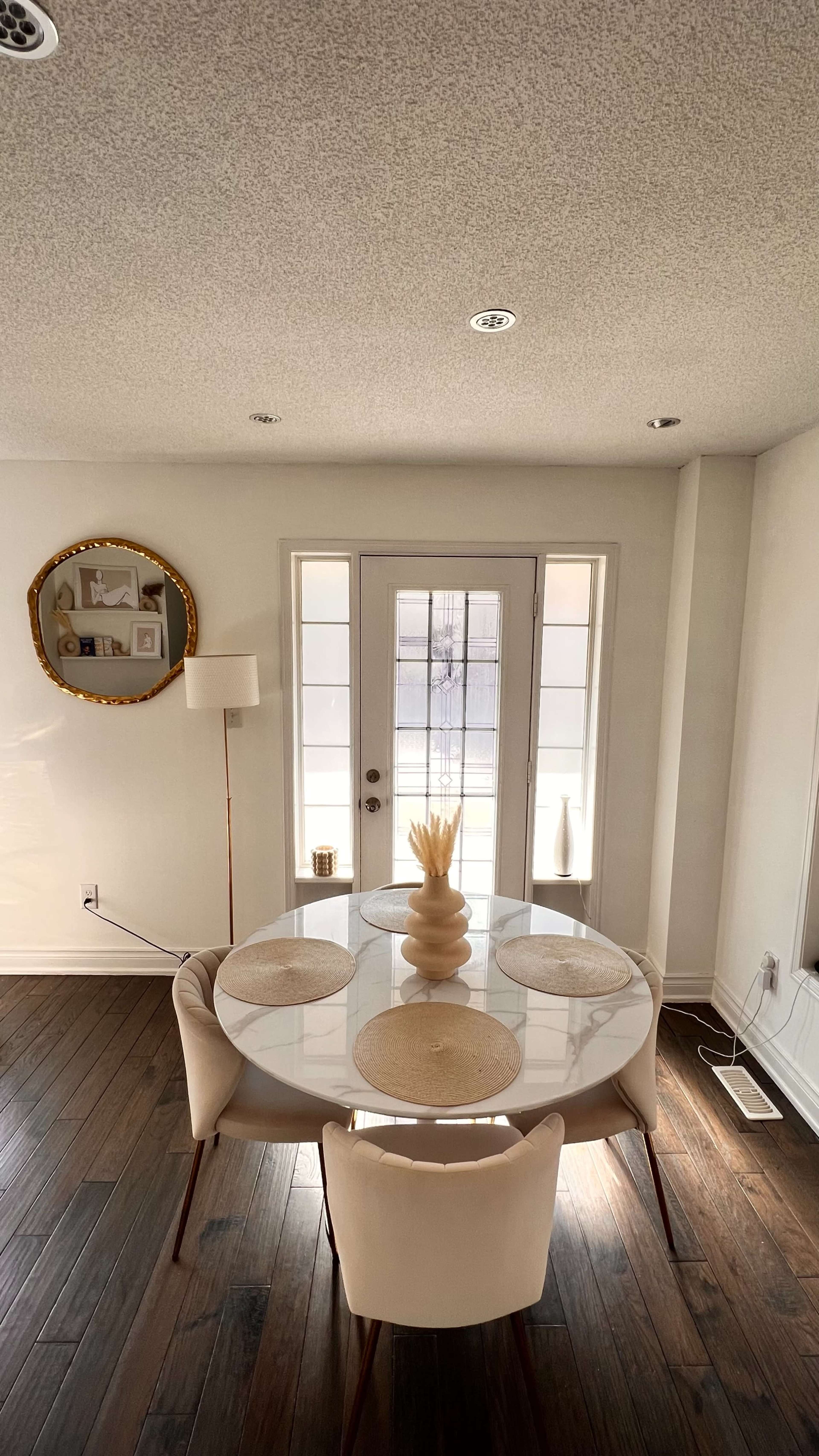 A modern dining area features a round marble table with four upholstered chairs, a floor lamp, and a decorative wall shelf near a pair of french doors.