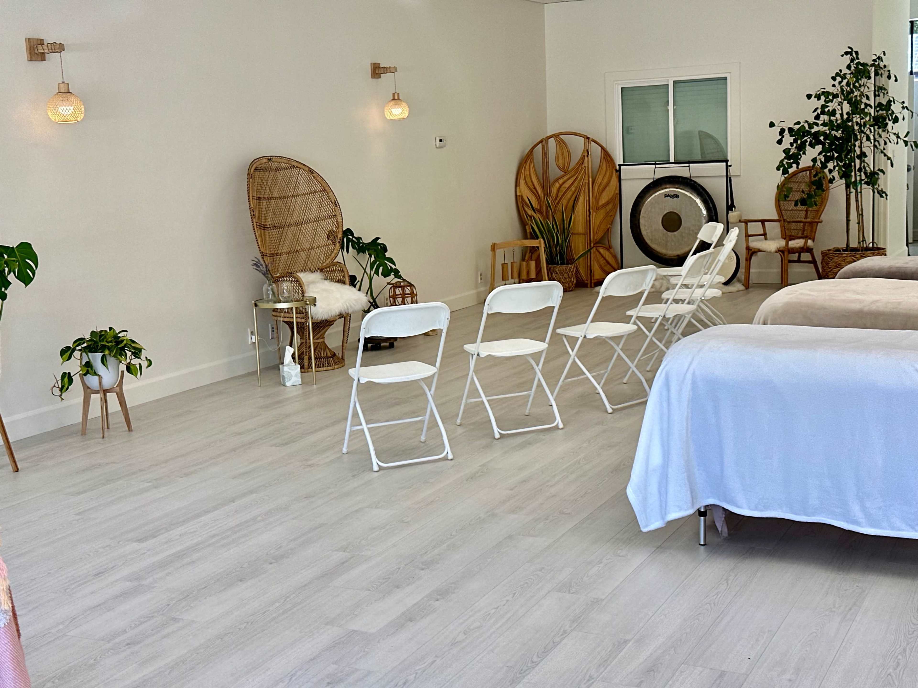 A serene wellness space featuring white folding chairs arranged in front of several massage tables, with decorative plants and woven furniture in the background.