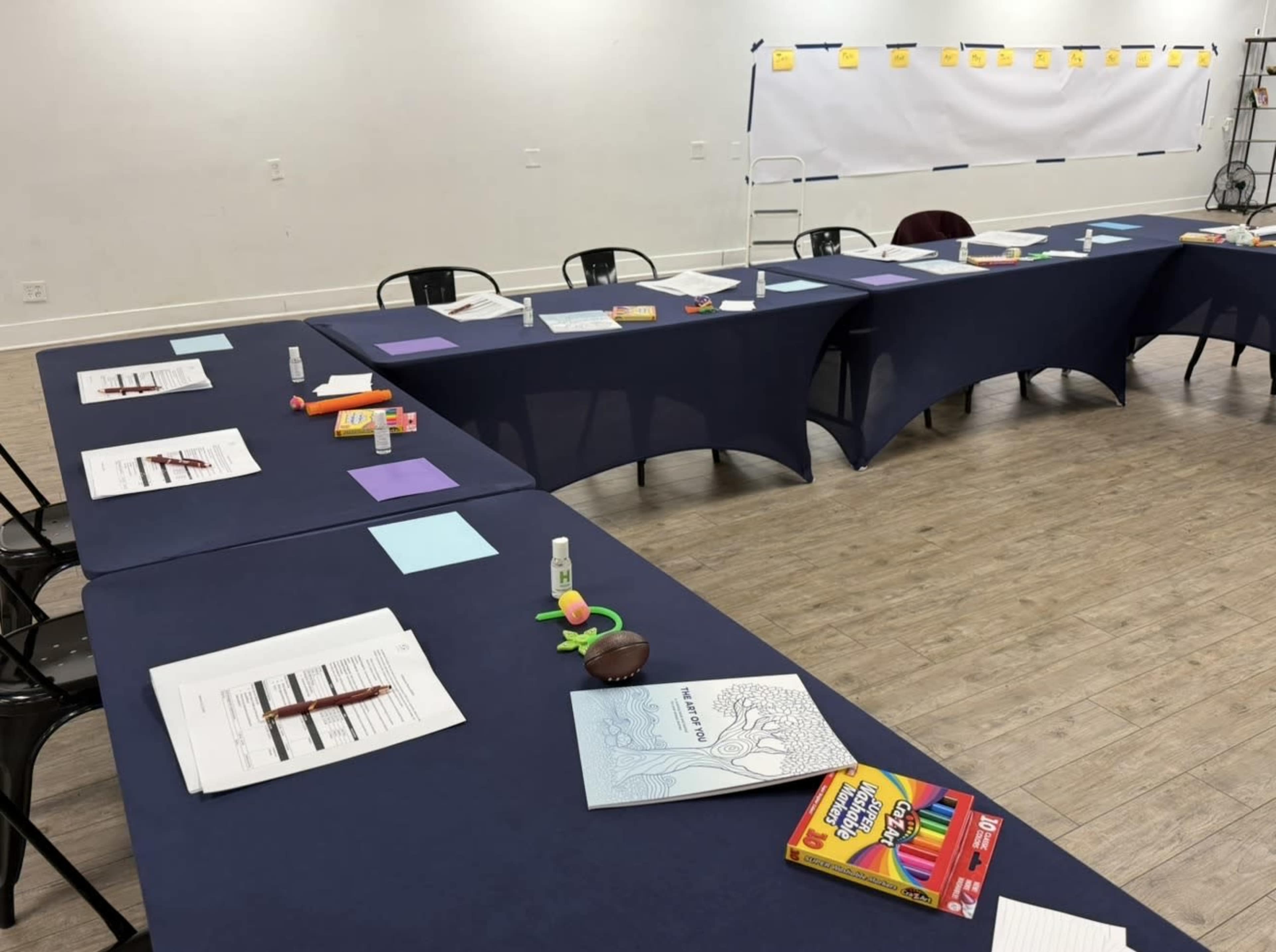 The image shows a classroom setting with several tables covered in dark blue cloths, each set up with printed materials, colorful writing supplies, and small artistic objects for a workshop or meeting.