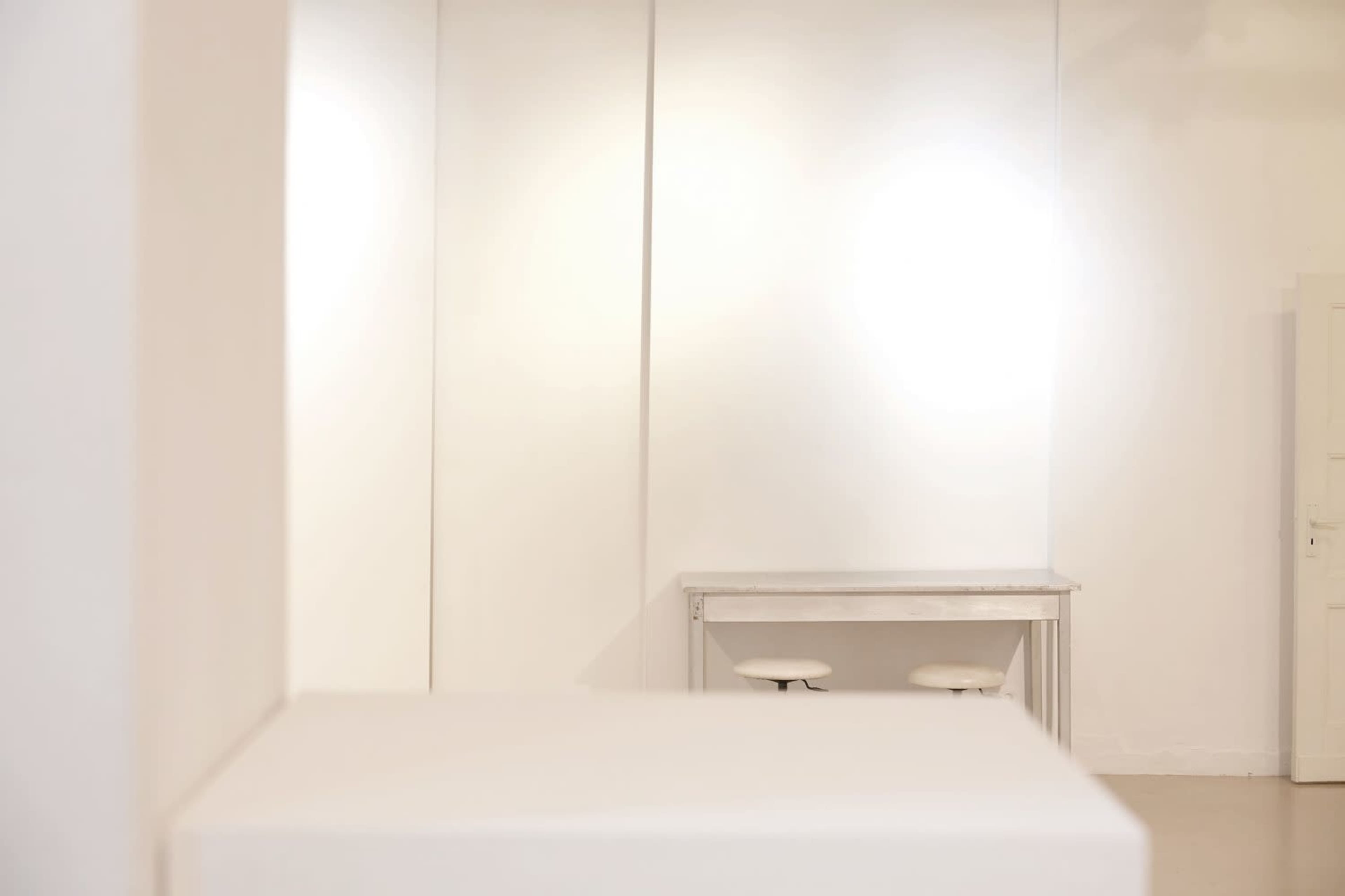 The image depicts a minimalist interior space with a white wall and a simple table with two stools.