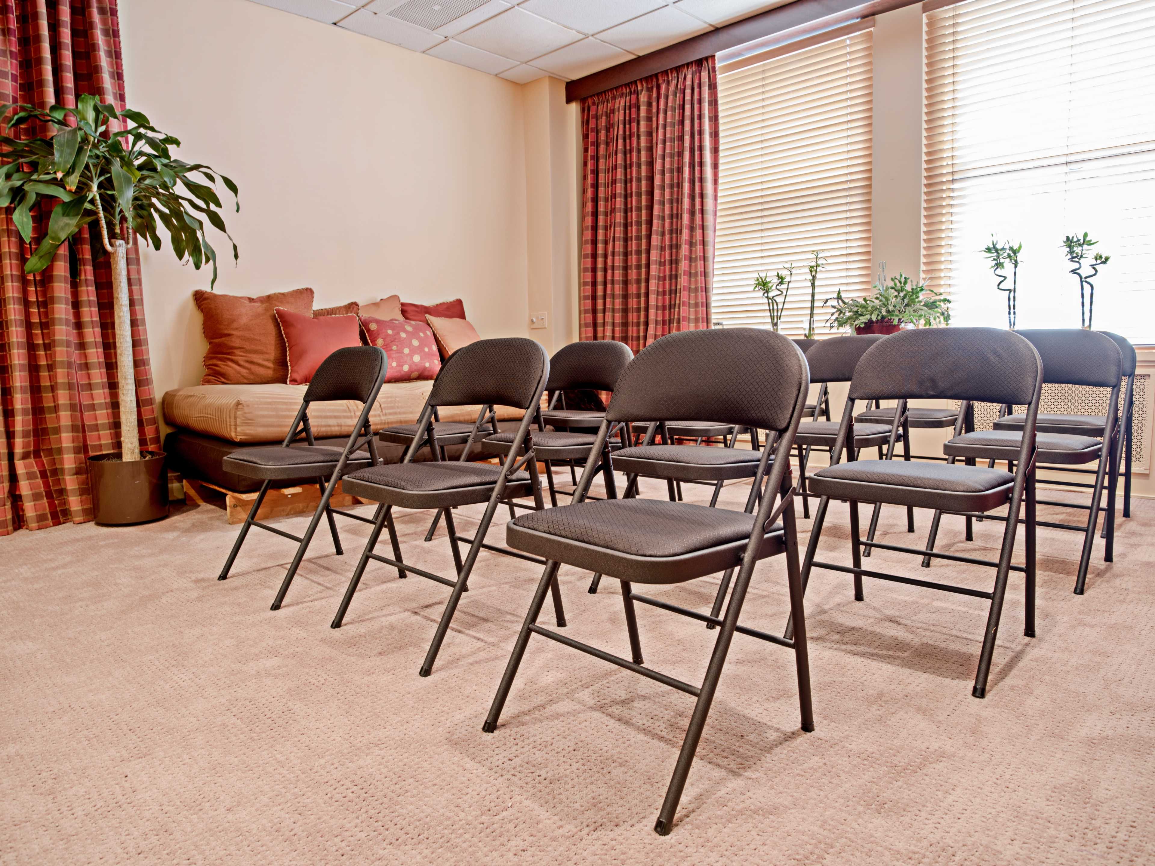 A row of black folding chairs is arranged in a meeting room with beige walls and large windows covered by striped curtains.
