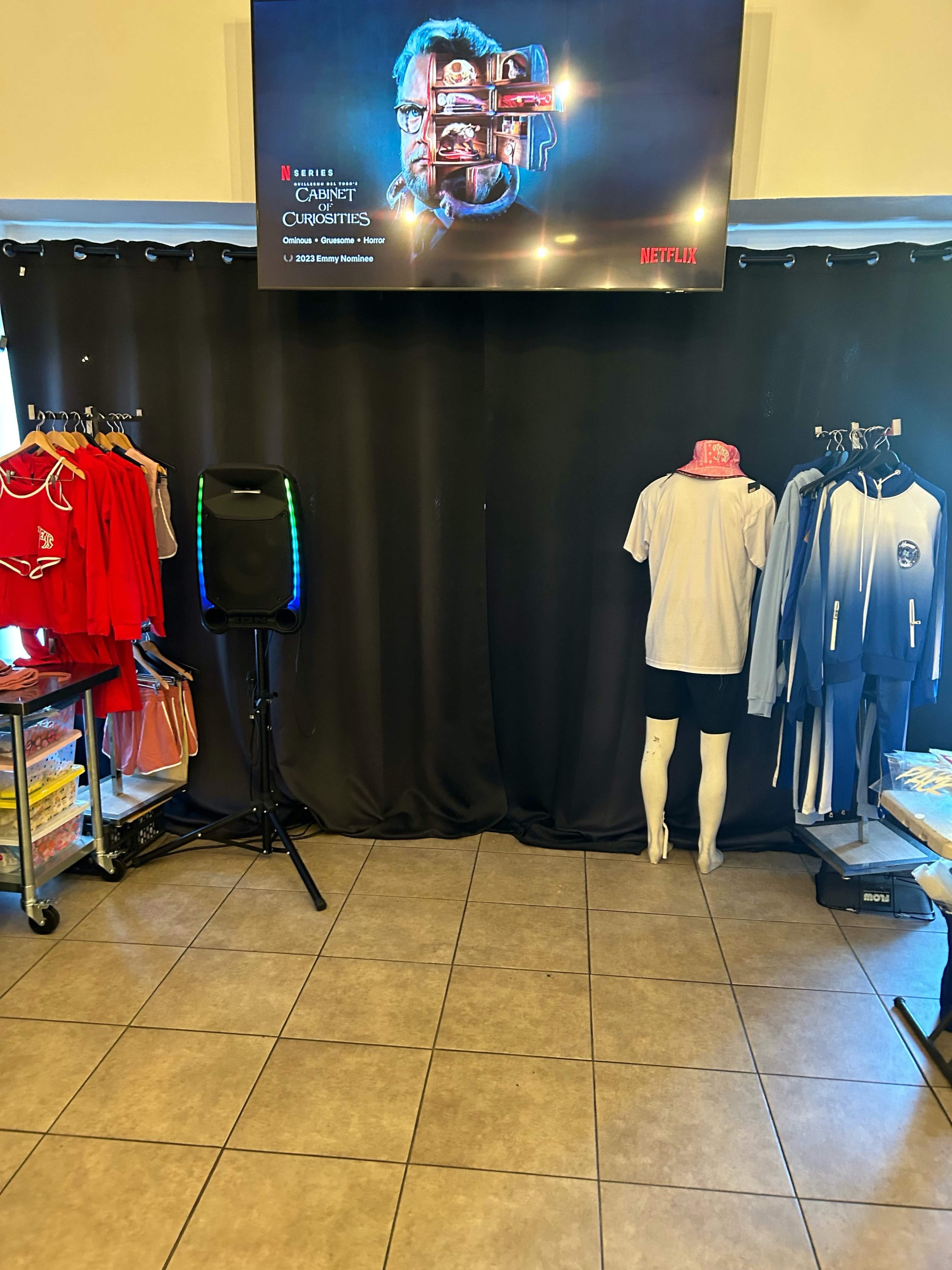 The image shows a retail space with clothing racks displaying various outfits, a speaker, and a large screen featuring promotional content, all situated in front of a black curtain.