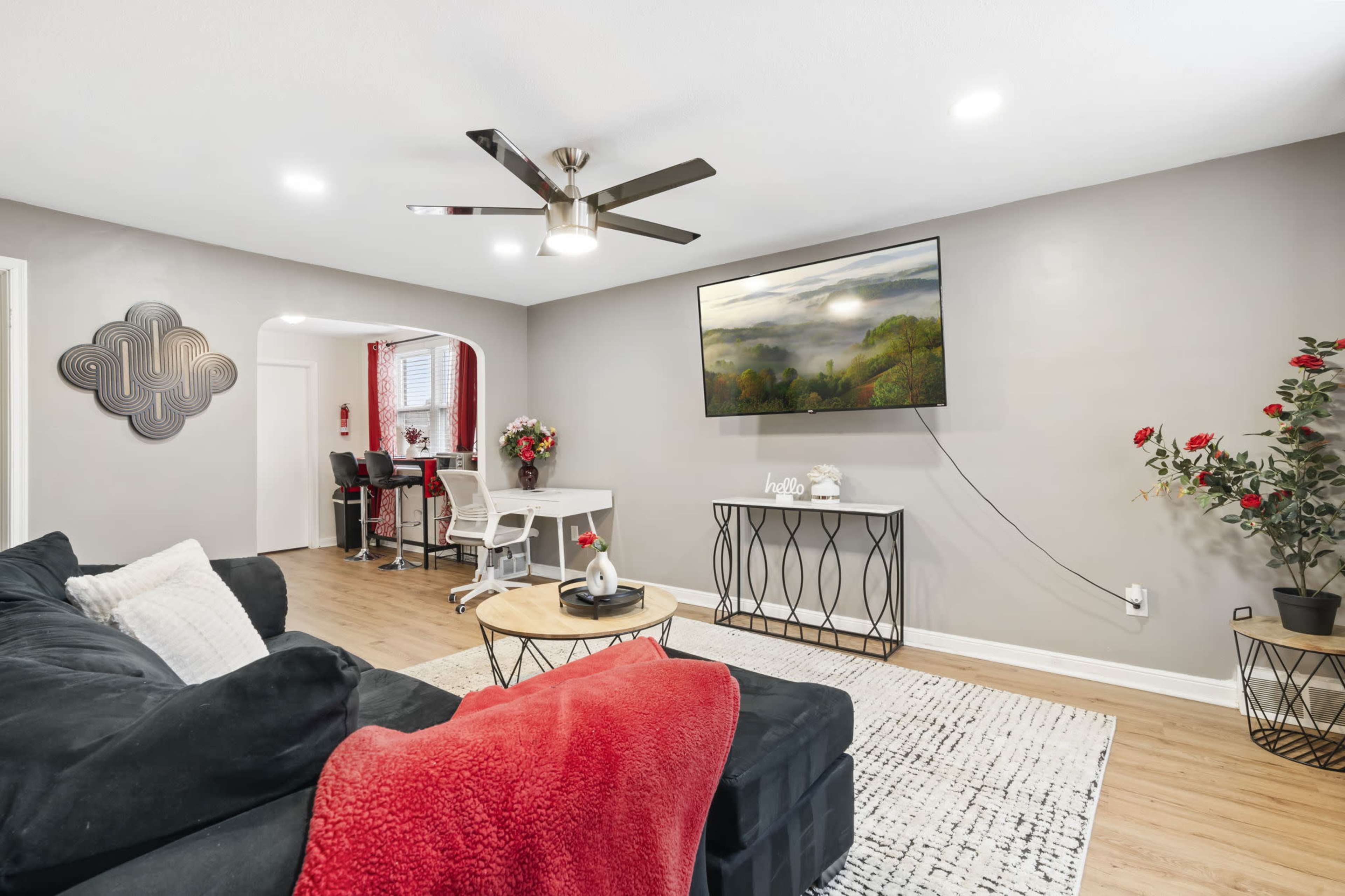 The living room features a dark sectional sofa with a red throw blanket, a wall-mounted television above a decorative console, and a dining area in the background.