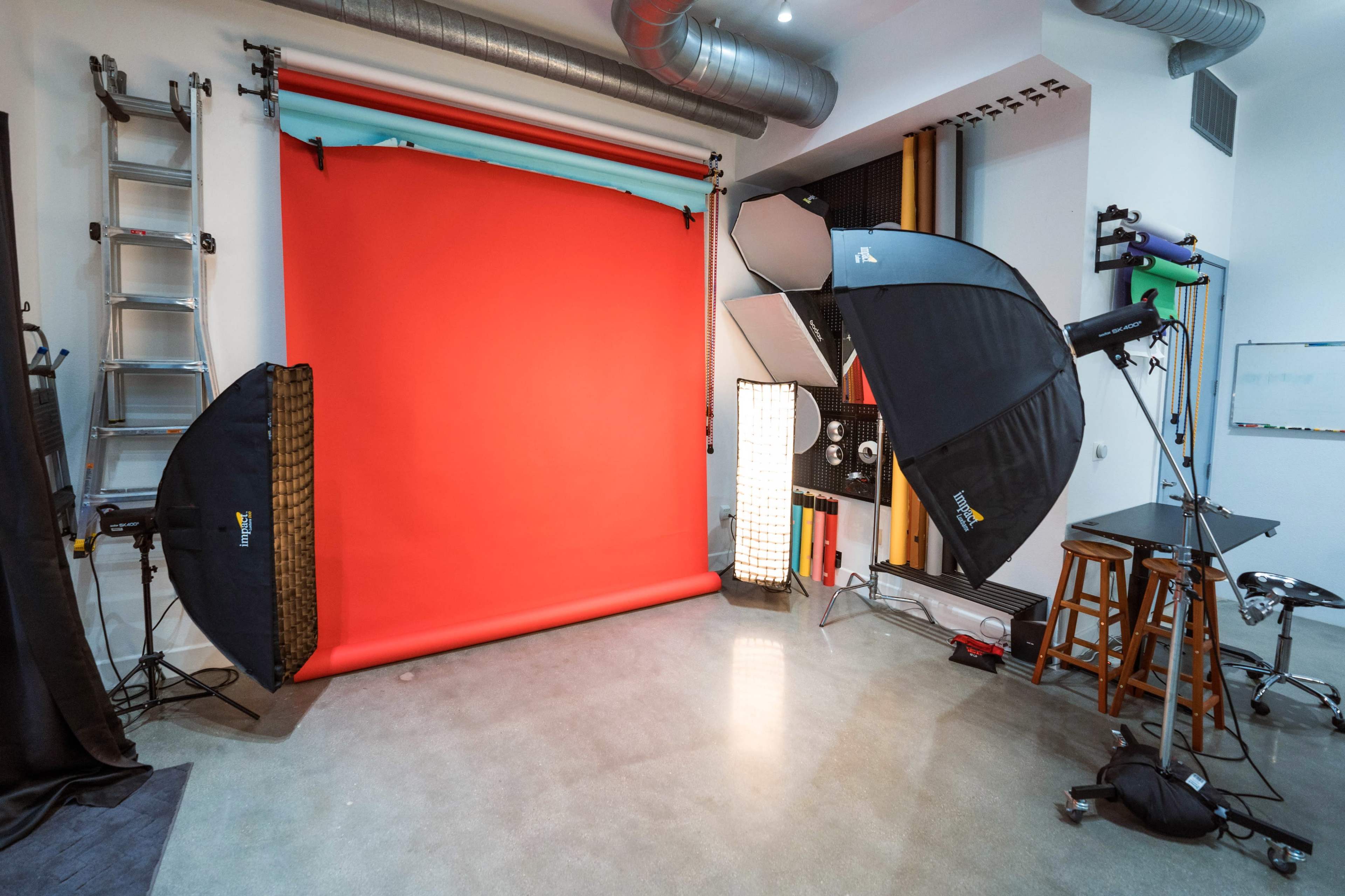A photography studio setup with a red backdrop, lighting equipment, a ladder, and various props arranged in the space.