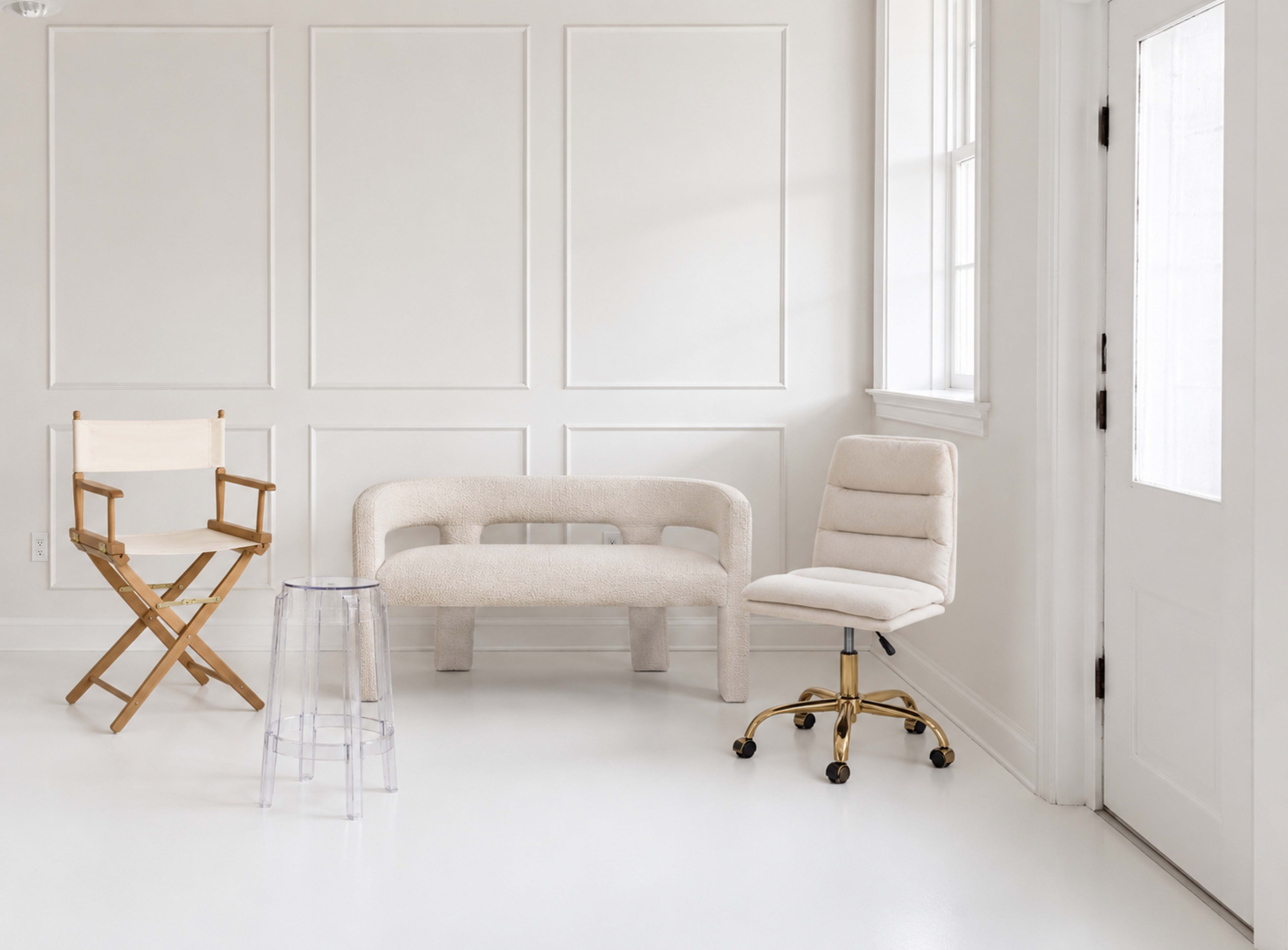 The image shows a minimalist interior with a white wall, a textured beige sofa, a clear acrylic table, a wooden director's chair, and a gold-accented office chair.
