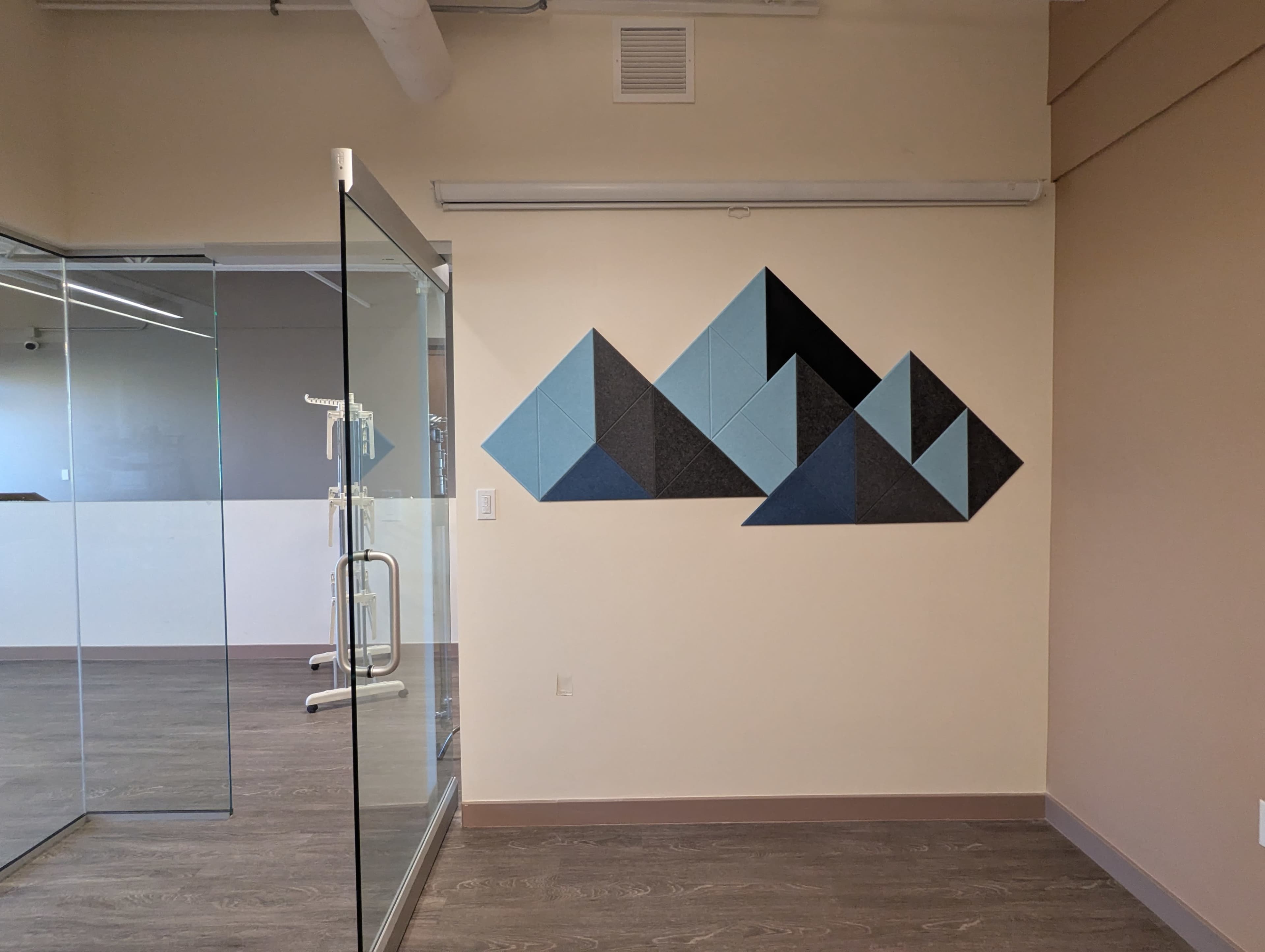 The image shows a wall in an office space featuring a geometric arrangement of felt panels in various shades of blue and black, adjacent to a glass partition.