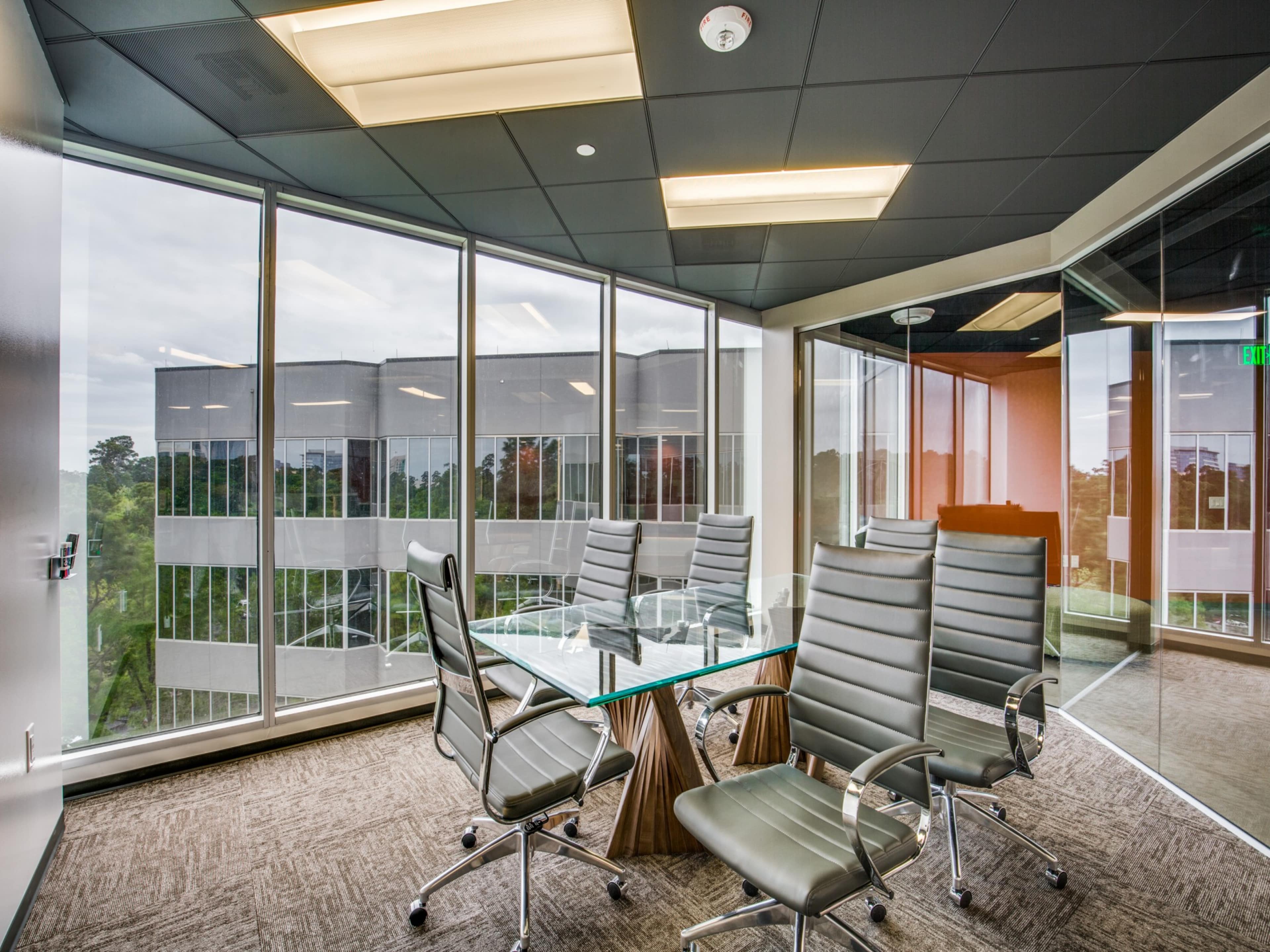 A modern conference room features a glass table surrounded by sleek chairs, with large windows overlooking a building and greenery outside.