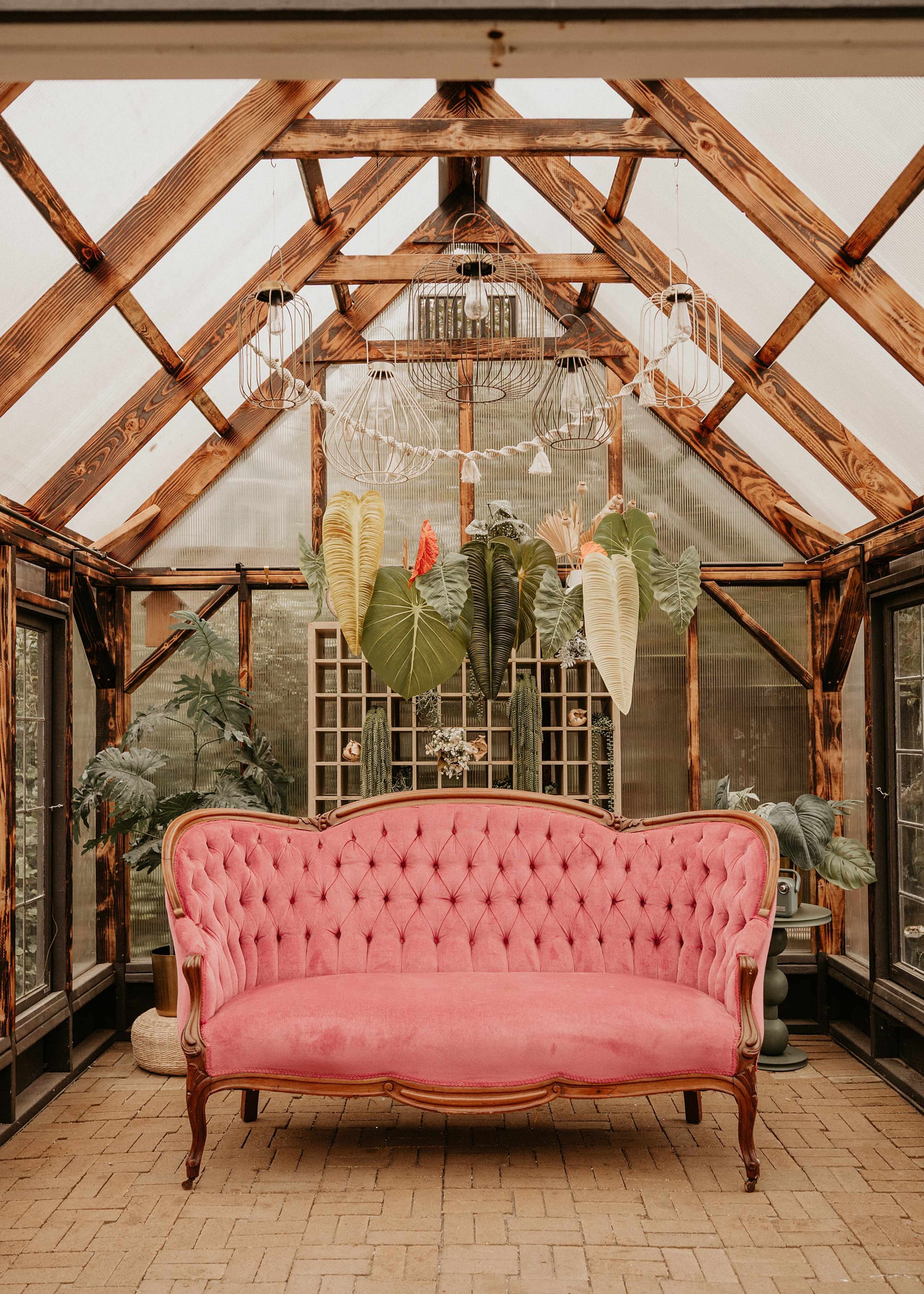 The image shows a pink tufted sofa positioned in a greenhouse filled with large plants and hanging light fixtures.