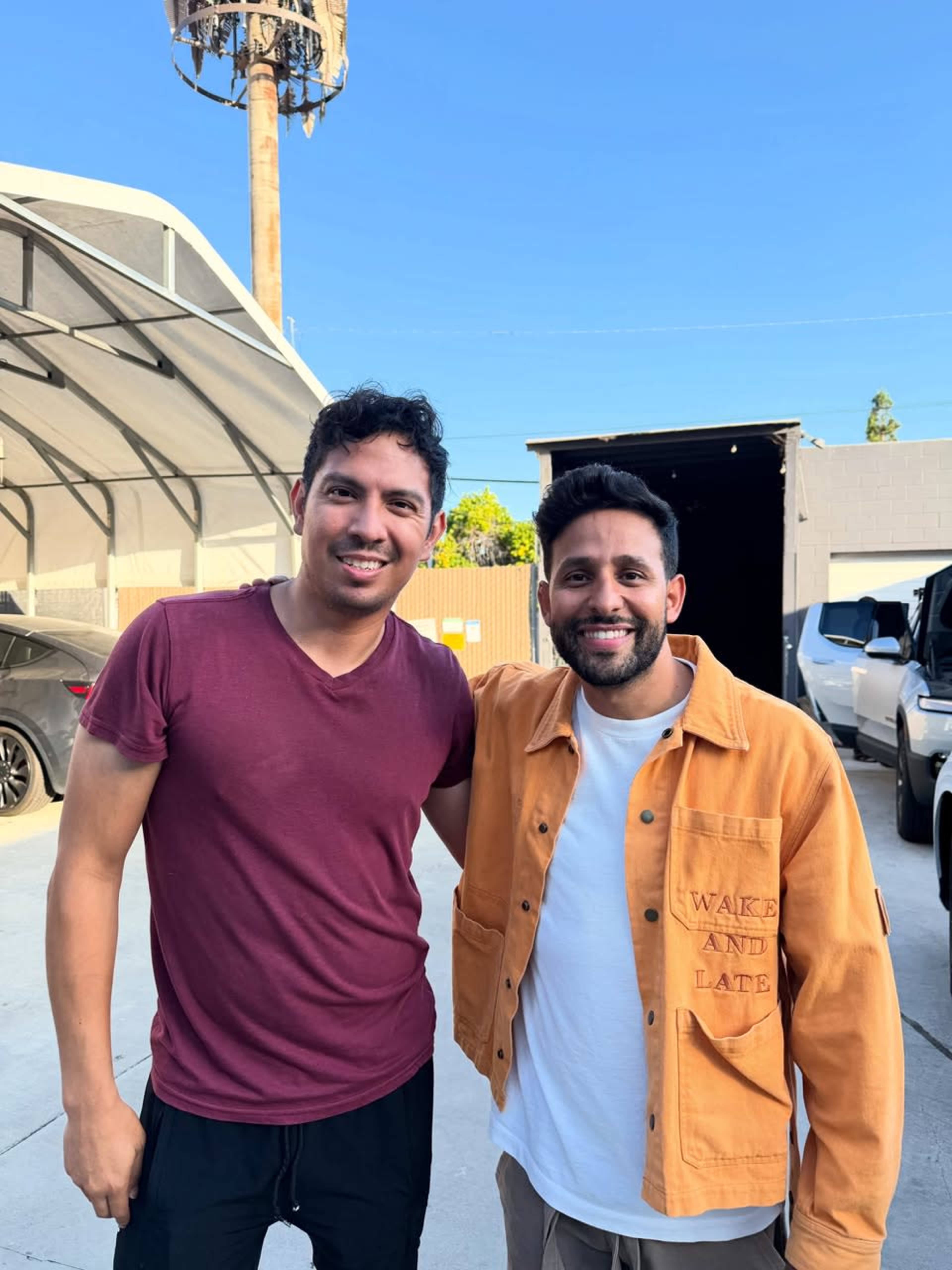 Two men are standing together, smiling in an outdoor setting with vehicles parked in the background.