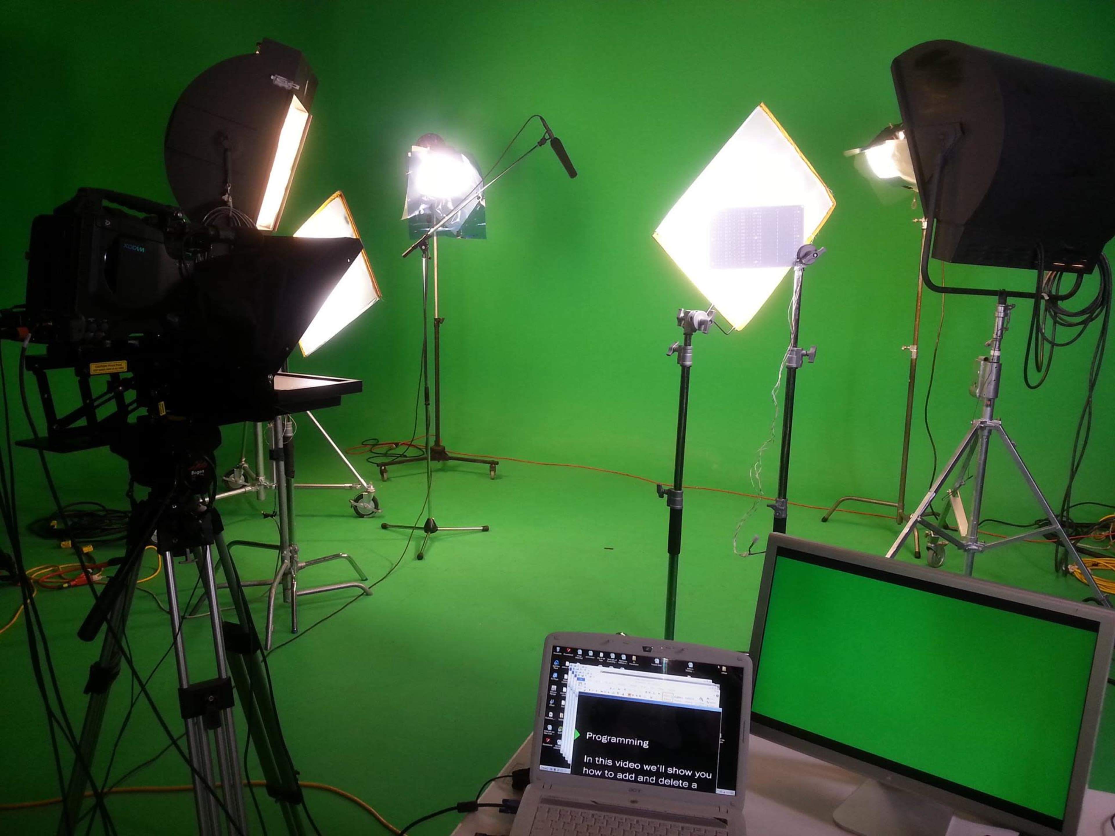 A green screen studio setup with multiple lights, a camera on a tripod, and a laptop displaying text.