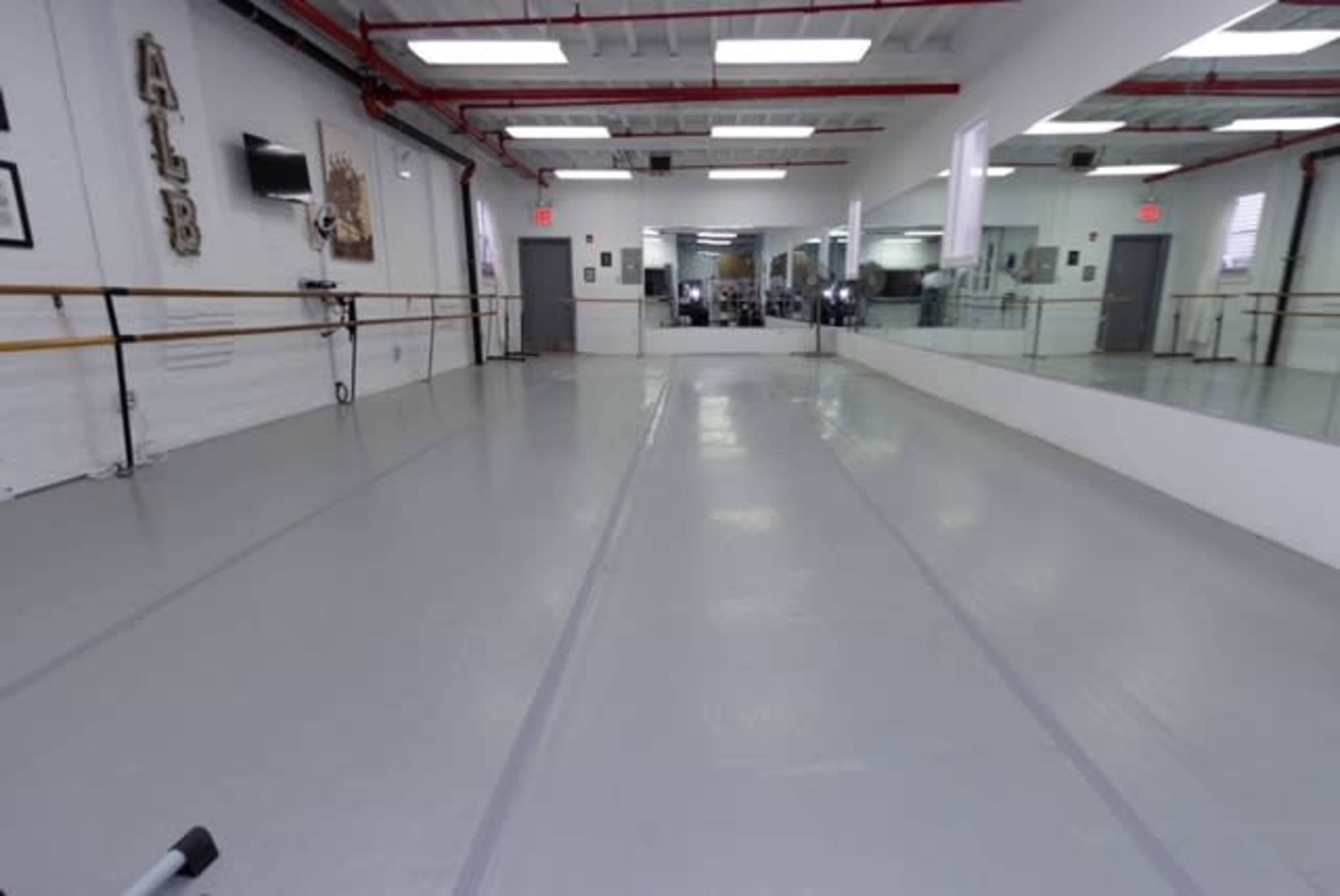 An empty dance studio with a mirrored wall, ballet bar, and gray flooring.