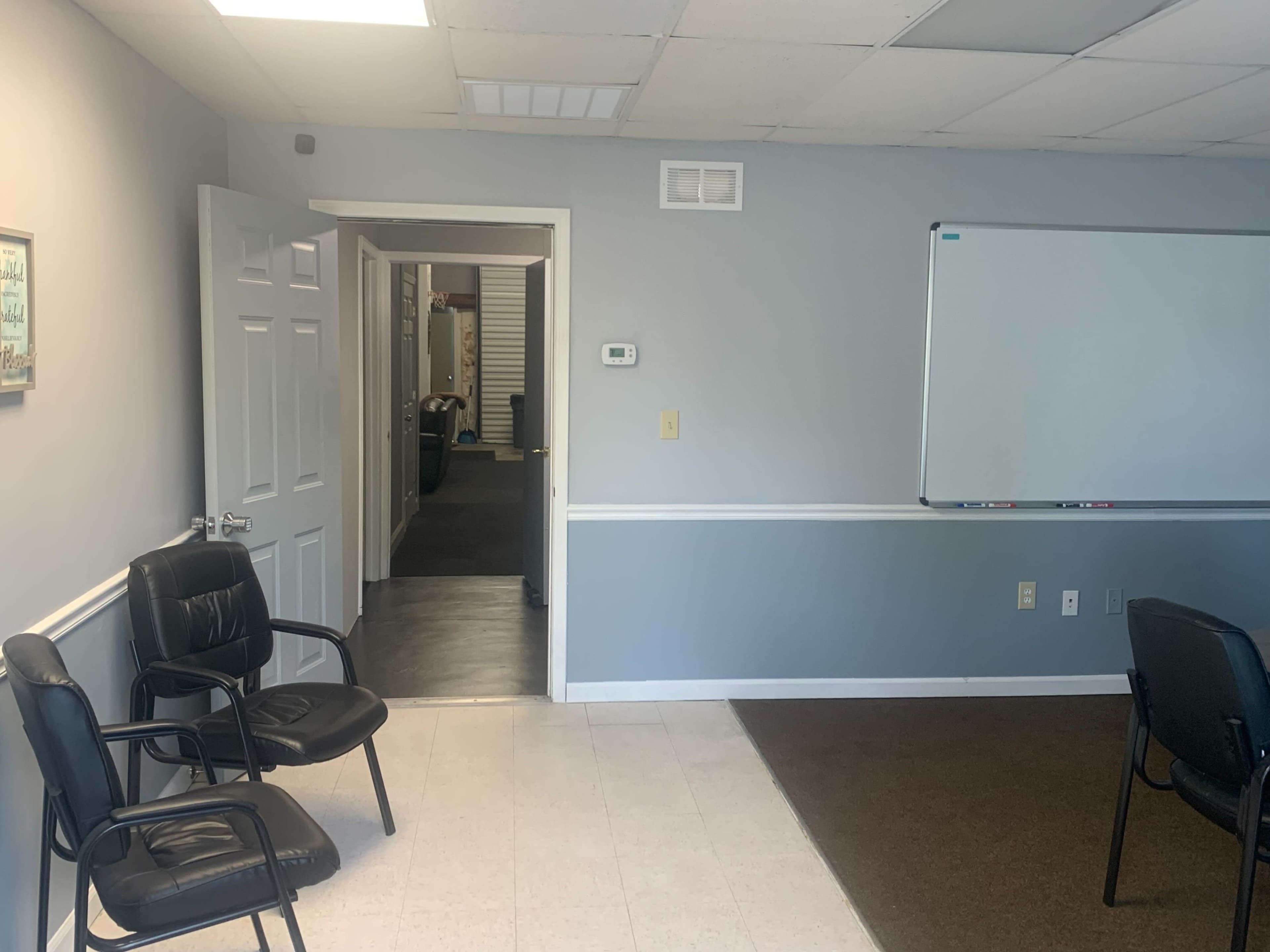 A conference room with two black chairs, a whiteboard on the wall, and an open door leading to a hallway.