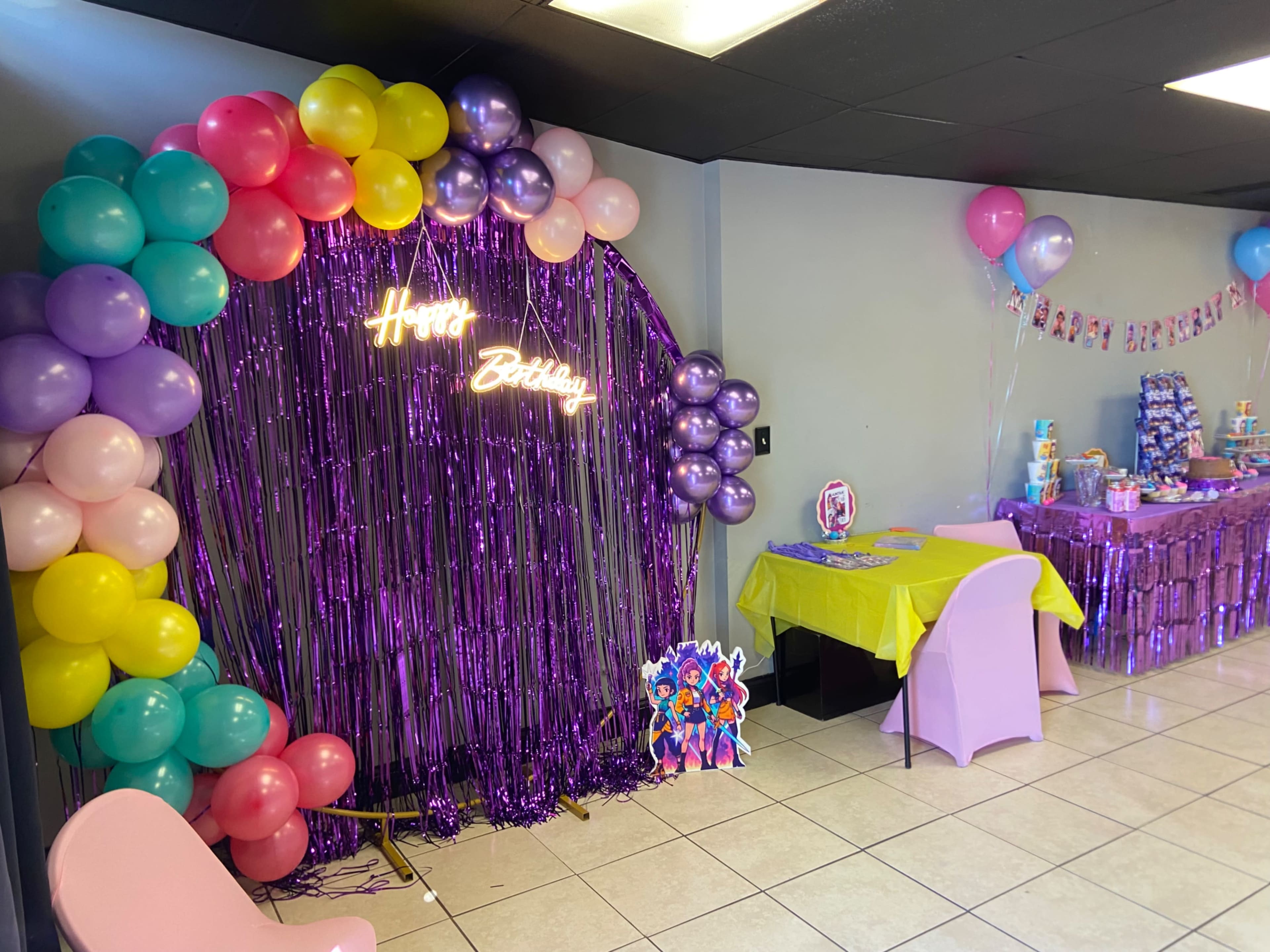 The image shows a birthday party setup featuring a colorful balloon arch, a decorated table with a cake, and various party decorations in a brightly lit room.